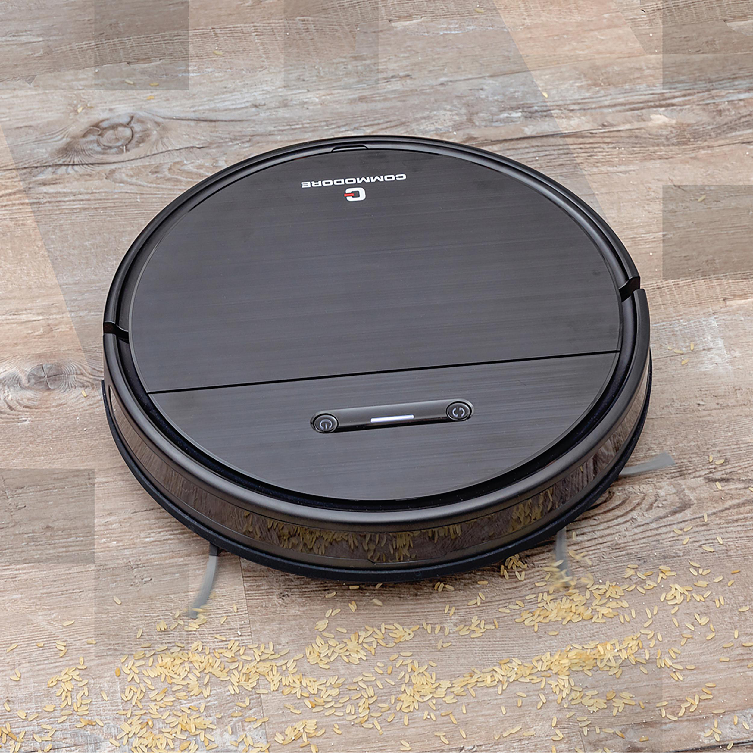 Schwarzer Commodore-Roboter-Staubsauger auf Holzboden, mit verstreuten Körnern.