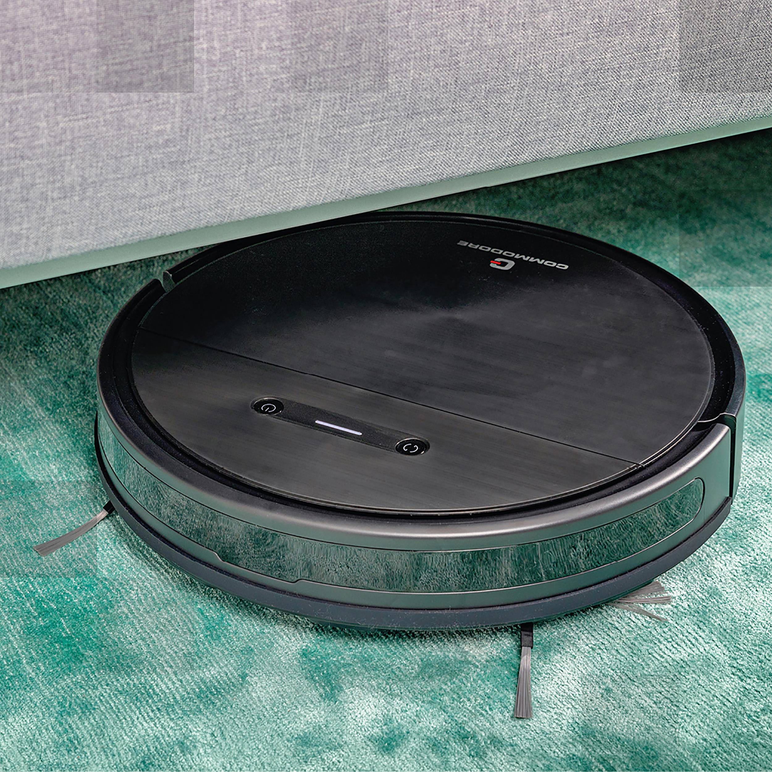Schwarzer Roboter-Staubsauger auf einem grünen Teppich neben einem grauen Sofabein.