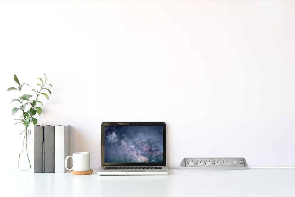Schreibtisch mit Laptop, der einen Nachthimmel zeigt, Büchern, Pflanze, Becher und eine Steckdosenleiste.