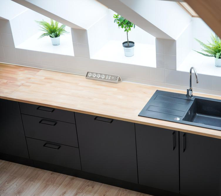 Moderne Küche. Schwarze Schränke, helle Holz-Arbeitsplatte. Spüle mit Wasserhahn. Fenster mit Topfpflanzen.