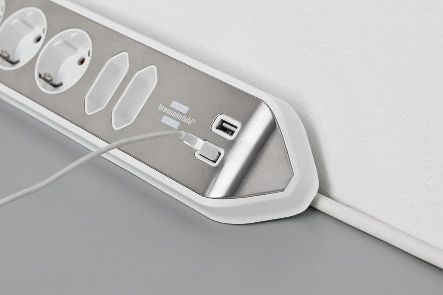 Eine weiße und silberne Steckdosenleiste, die in eine graue Oberfläche eingesteckt ist, mit einem Ladekabel im USB-Anschluss.