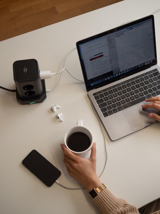 Eine Person arbeitet am Laptop, mit Telefon, Kaffee und Netzteil.