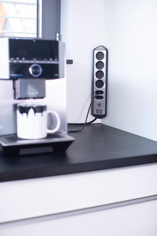 Kaffeemaschine neben einer Steckdosenleiste auf einer schwarzen Arbeitsplatte, mit weißer Wand.