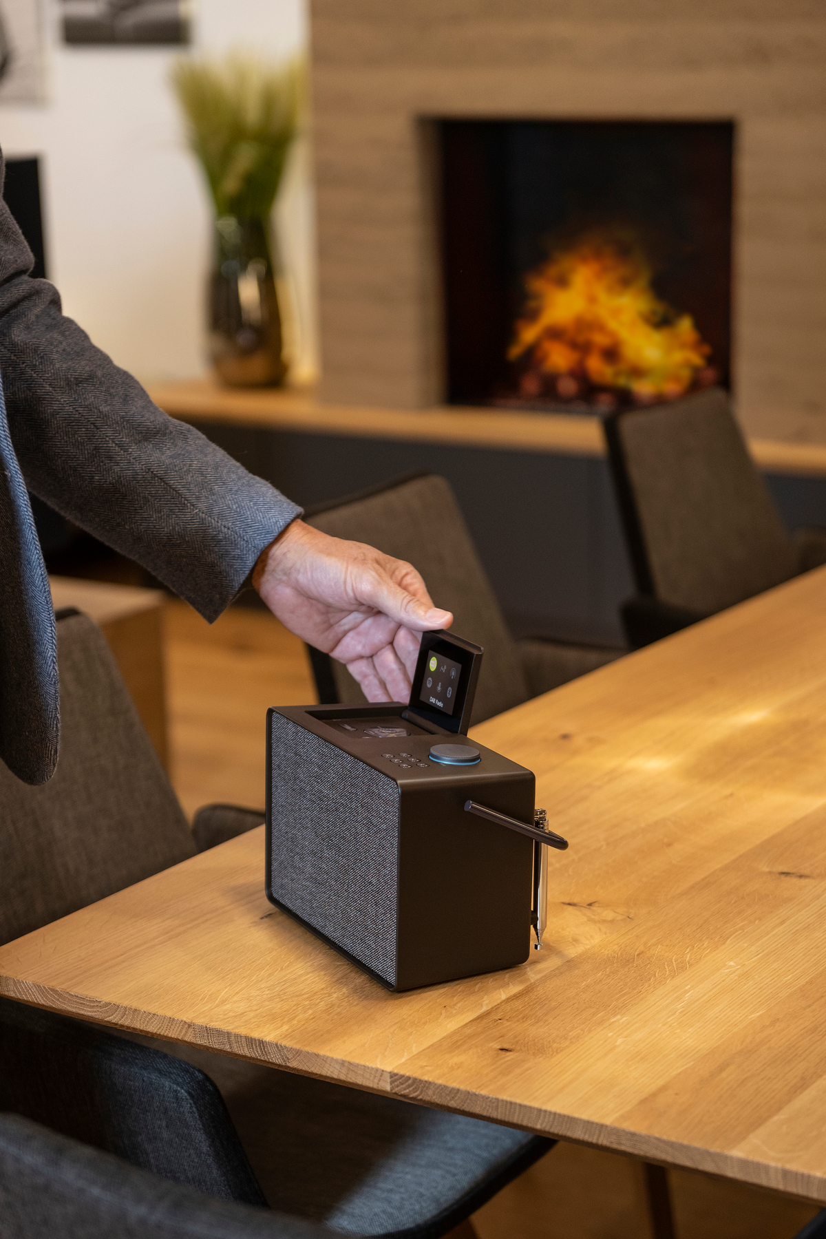 Une main tient une petite radio noire avec un écran et une table en bois devant une cheminée.