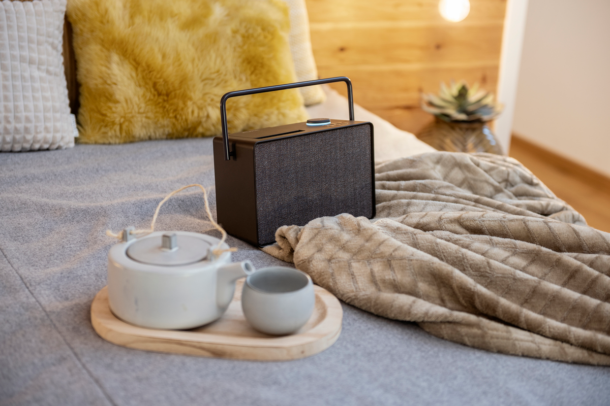 Un haut-parleur noir est sur un lit avec un plateau et un service à thé. Une couverture et des oreillers beiges sont présents.