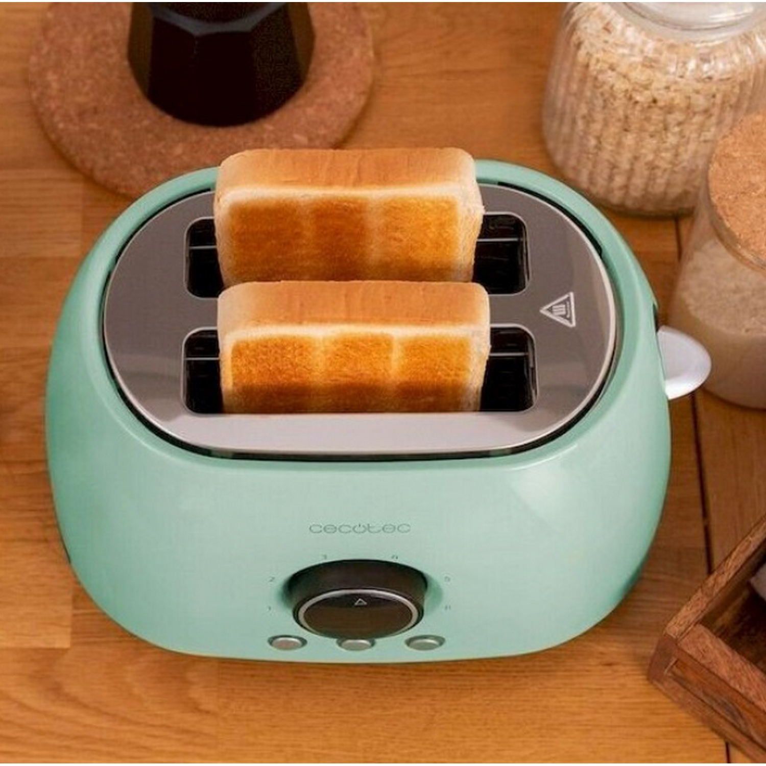 Una tostadora verde menta con dos rebanadas de pan tostado. Otros elementos en una mesa de madera incluyen una cafetera y frascos.