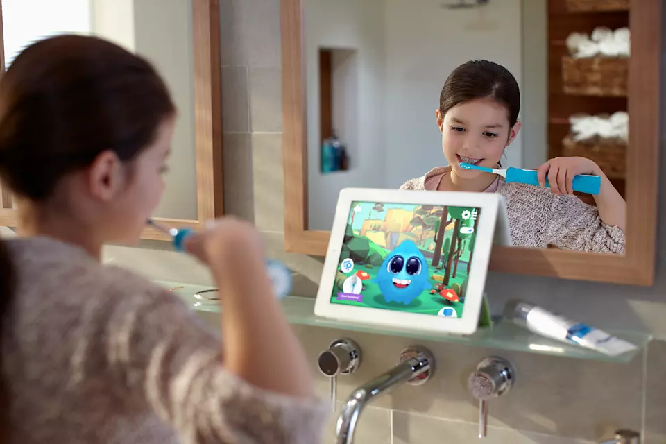 Une fille se brossant les dents devant un miroir, regardant une tablette avec un dessin animé.