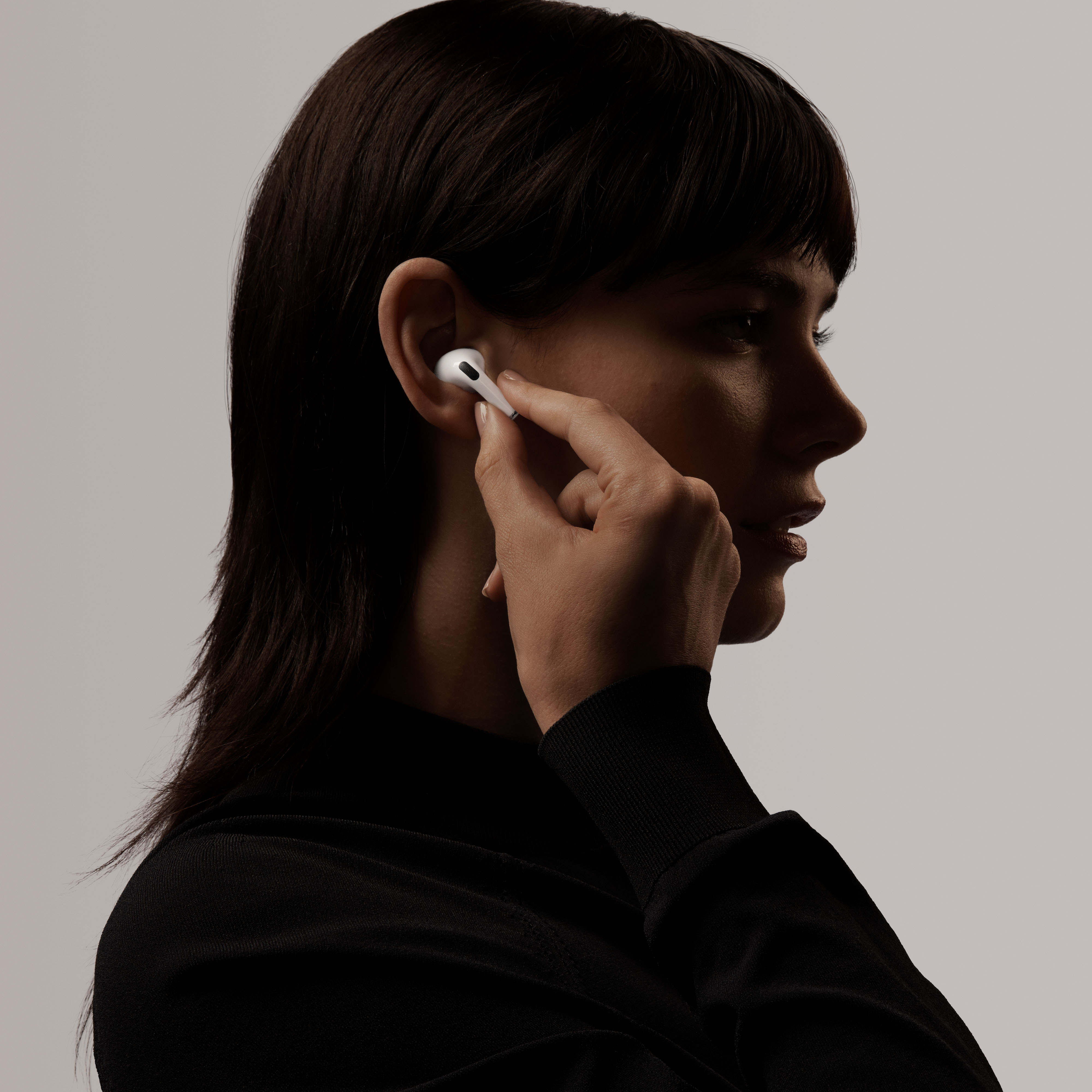 Siyah bir üst giyen, beyaz bir AirPod Pro'yu kulağında tutan, degrade bir arka planı olan bir stüdyoda bir kadın.