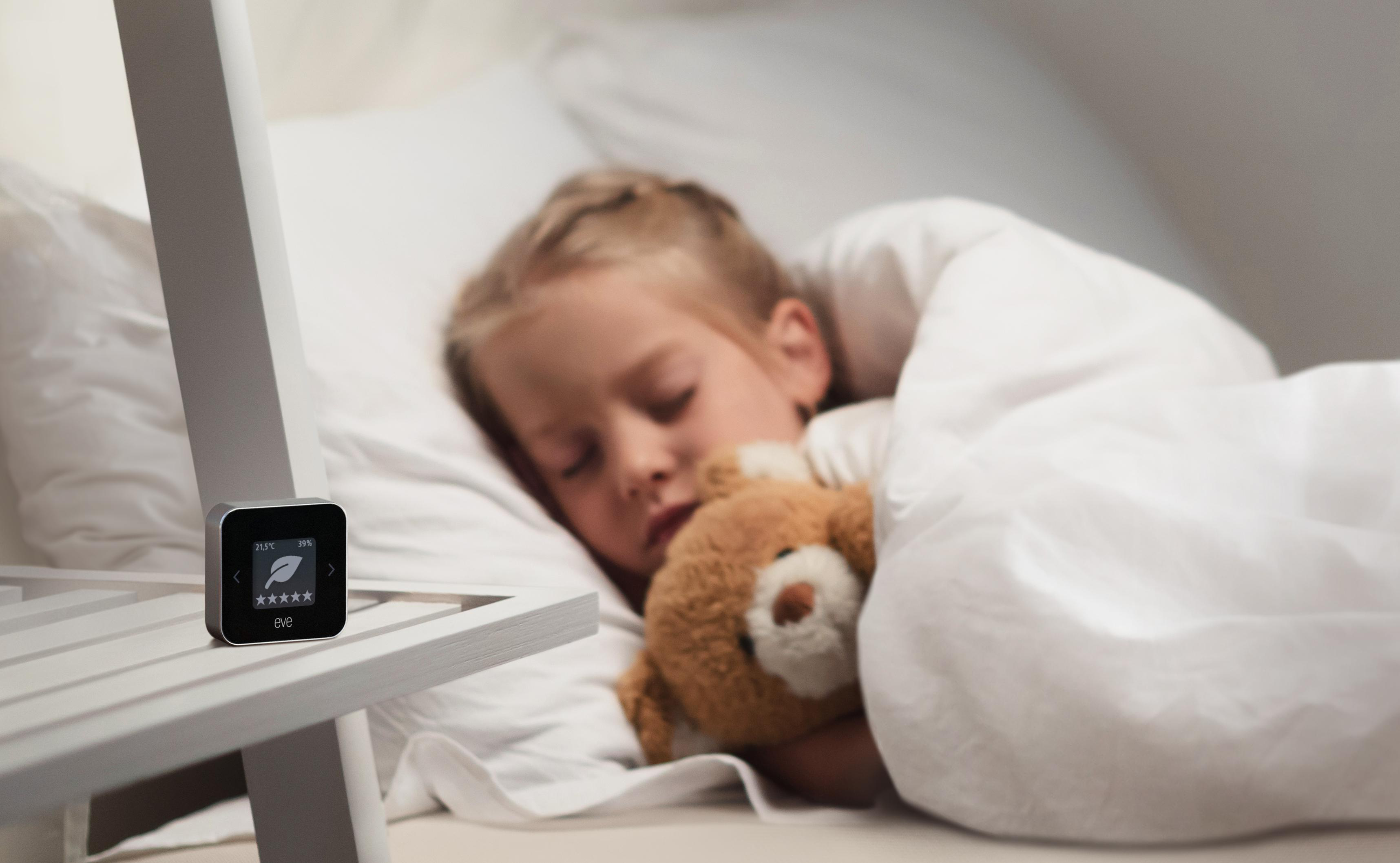 Ein schlafendes Kind mit Teddybär, neben einem digitalen Thermometer auf einem Nachttisch.