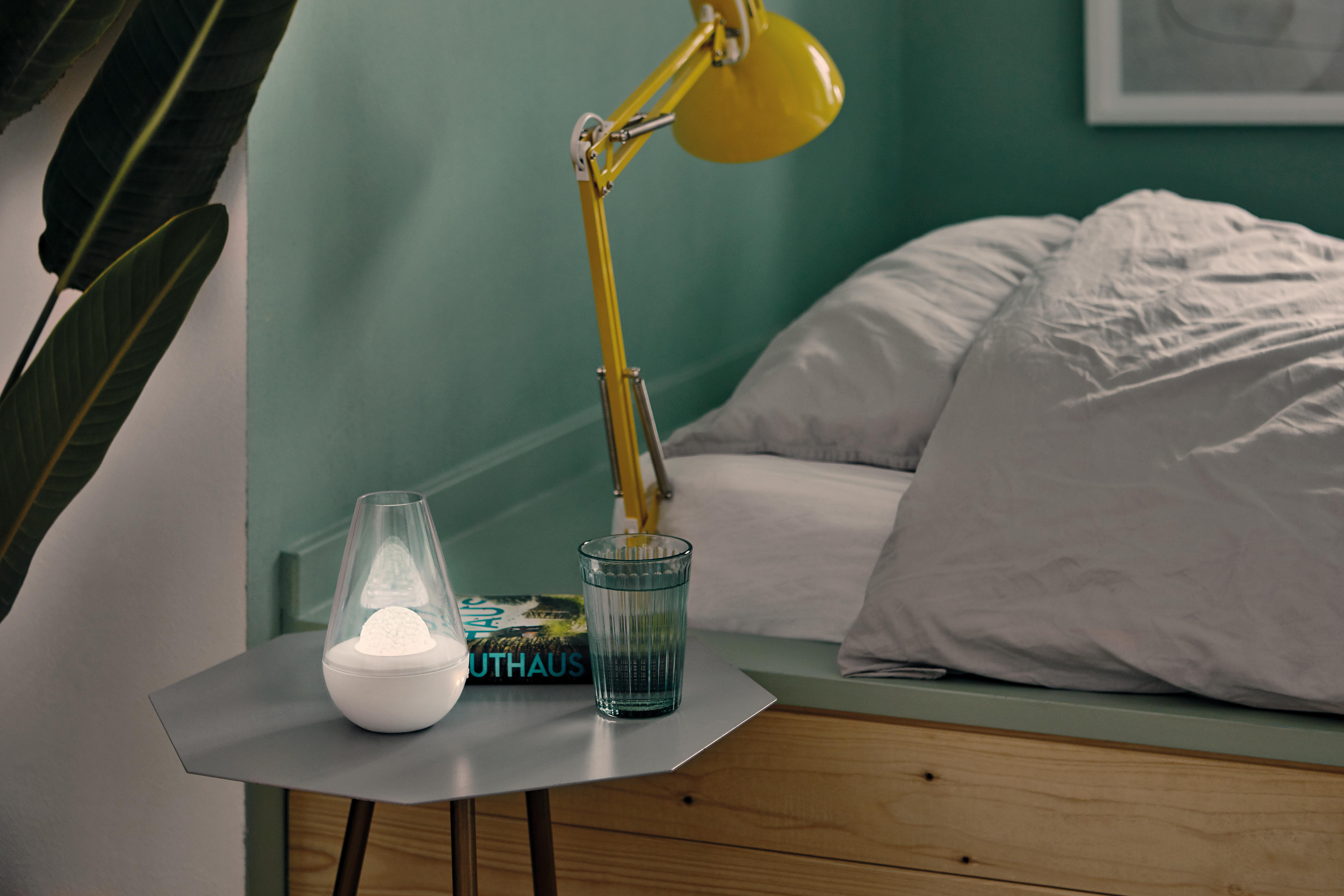 Eine Schlafzene mit Tisch, Lampe und einem Glas Wasser. Bett und grüne Wand im Hintergrund.