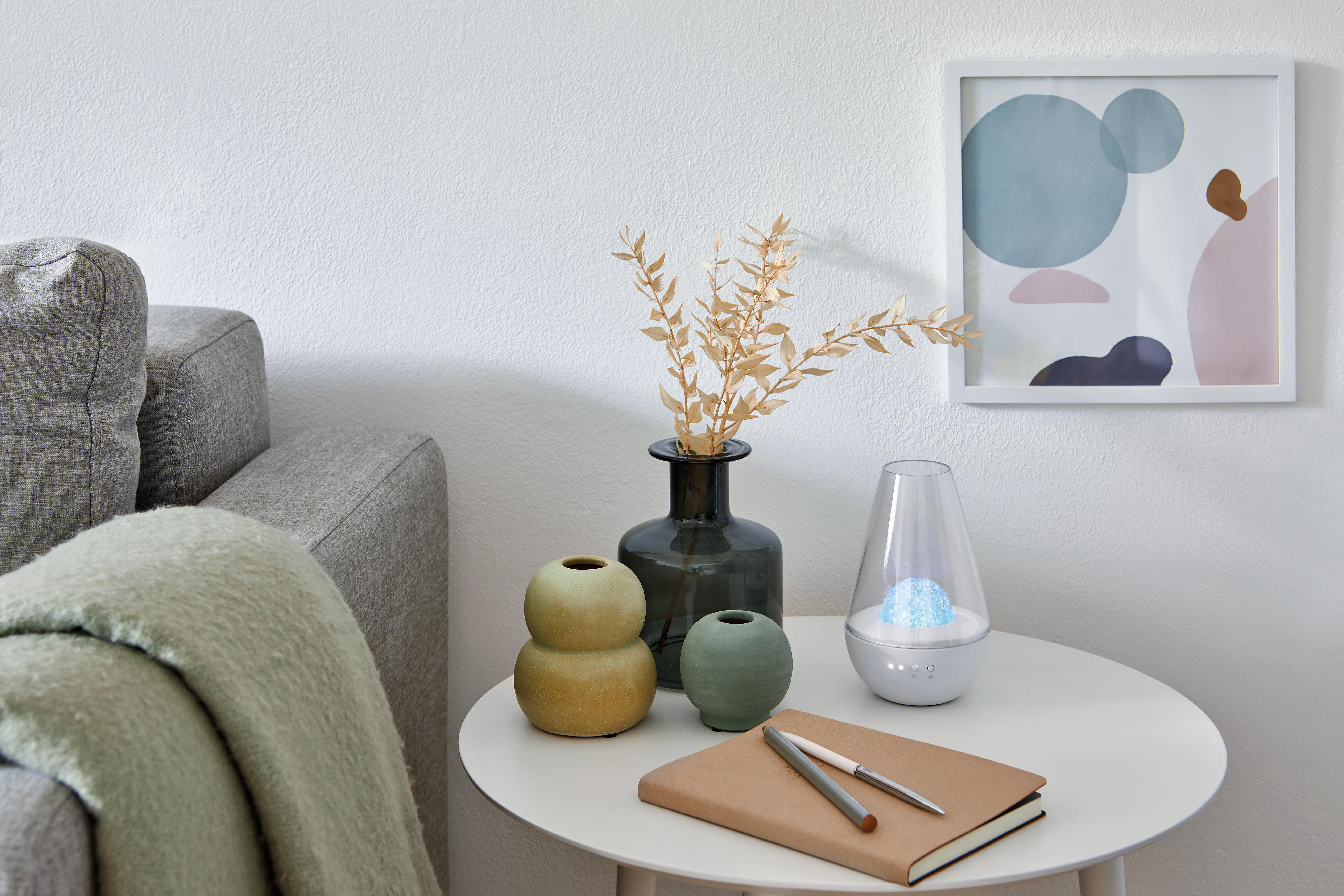 Ein Tisch mit Vase, getrockneten Pflanzen und Lampe. Notizbuch und Stifte sind vorhanden, sowie ein Bild an der Wand.