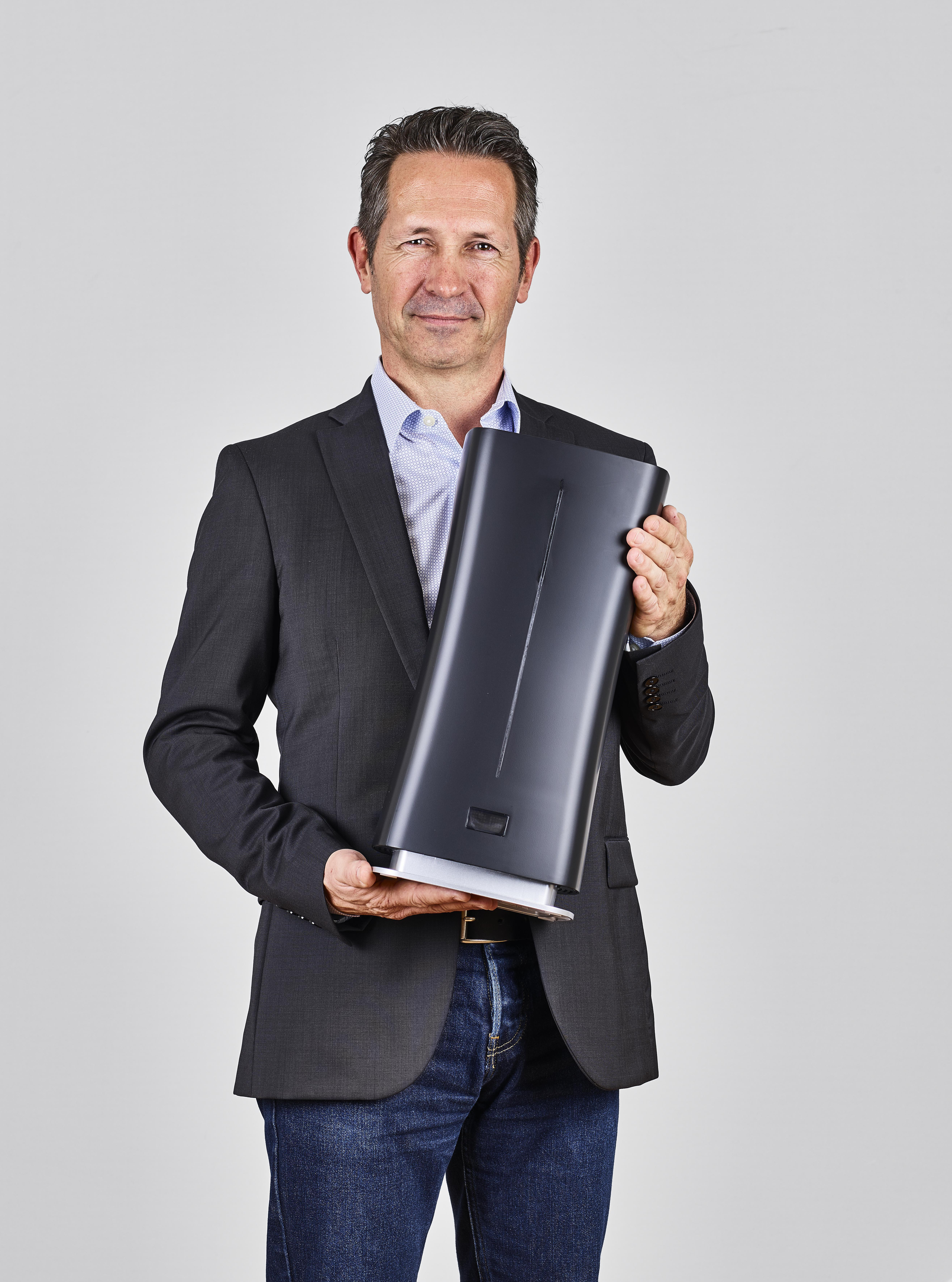 Ein Mann hält ein dunkles, schlankes Gerät, trägt Anzug und Jeans, vor weißem Hintergrund.