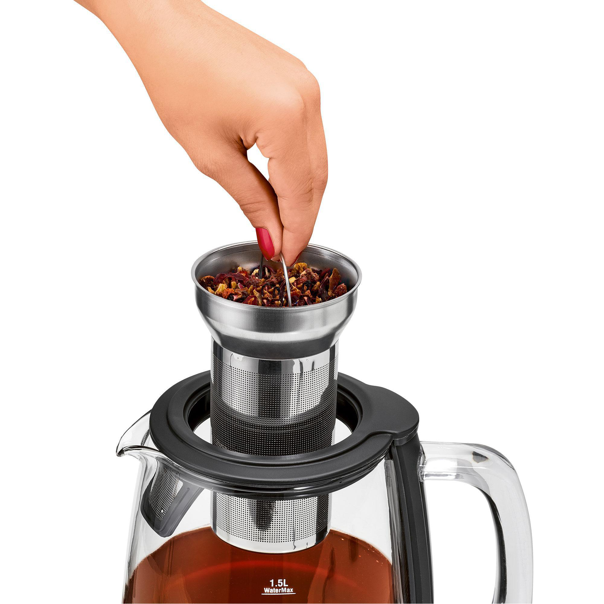 Eine Hand legt Teeblätter in einen silbernen Tee-Infuser in einer gläsernen Teekanne.