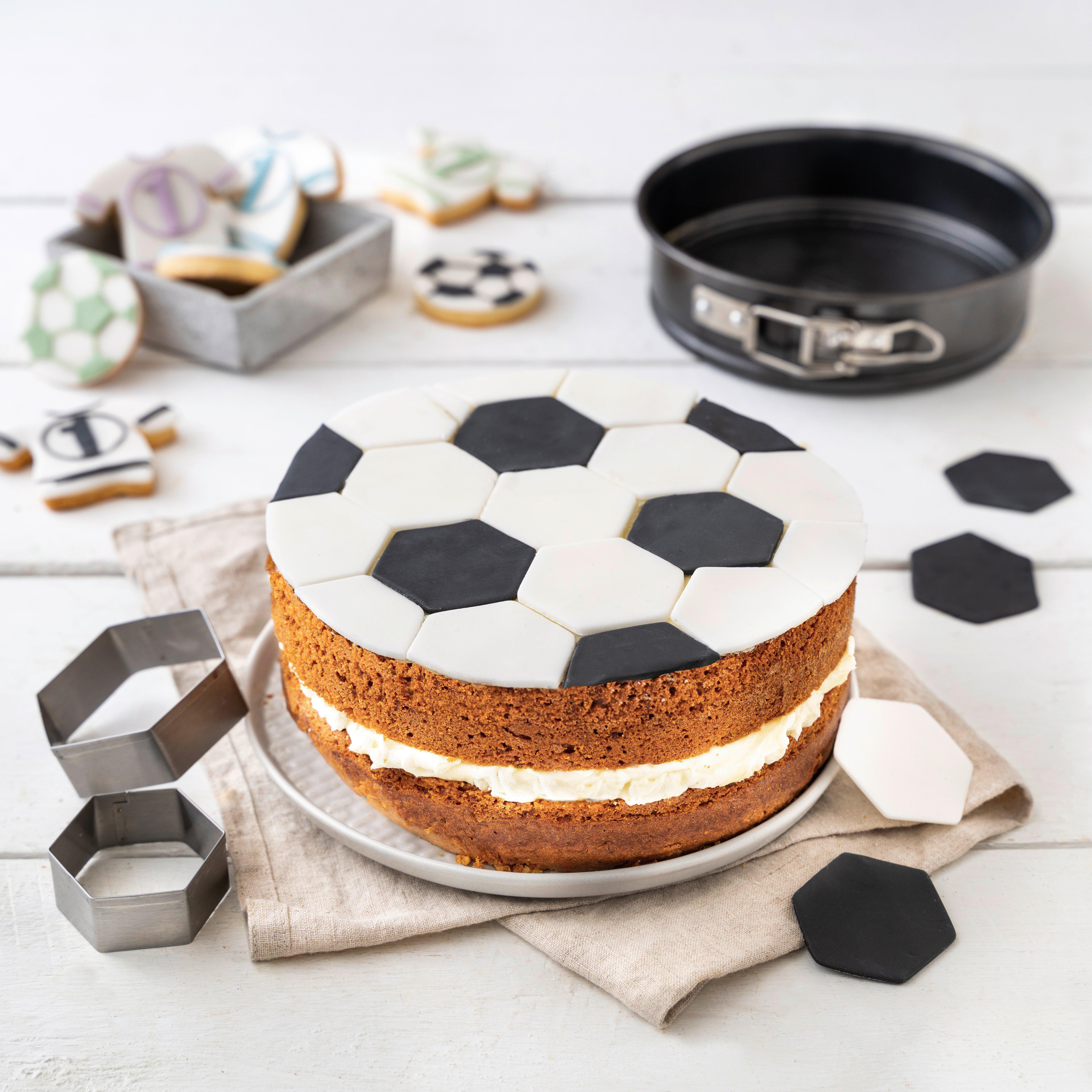 Ein Fussballkuchen, dekoriert mit schwarz-weißen Sechsecken. Backwerkzeuge und Kekse im Hintergrund.
