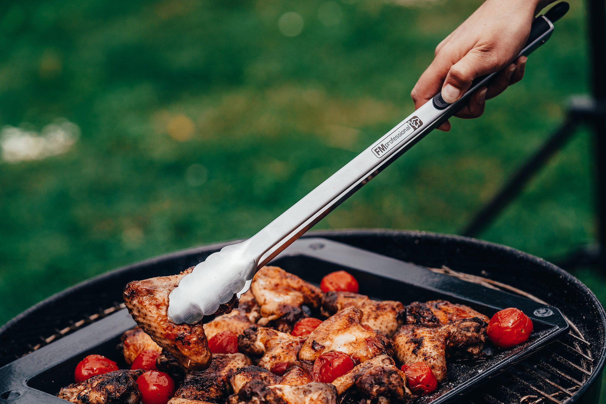 Person grillt Flügel und Tomaten mit Zangen auf einem Grill.