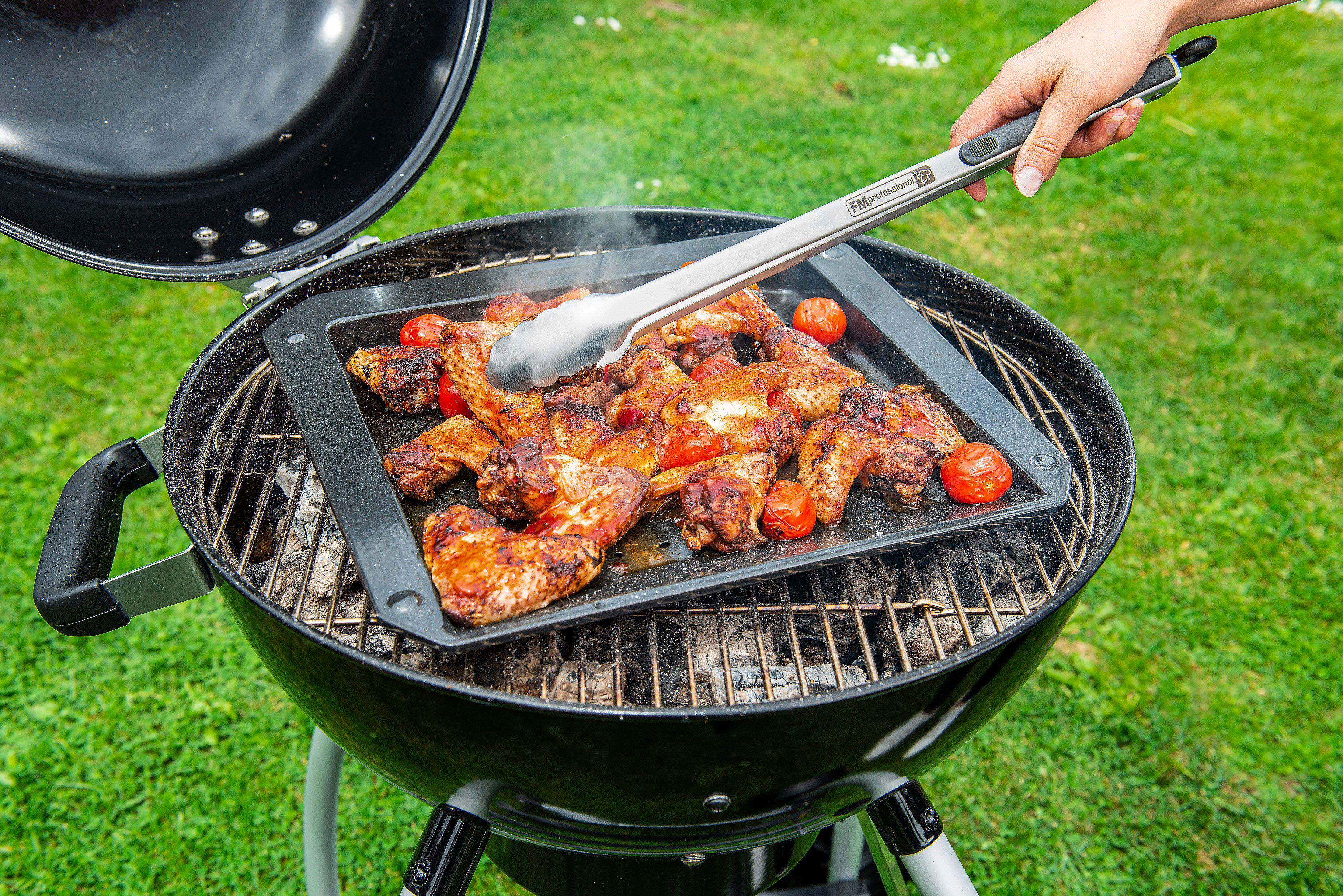Grill mit Hähnchenflügeln, Tomaten und Zangen. Person benutzt Zangen. Grüner Hintergrund.