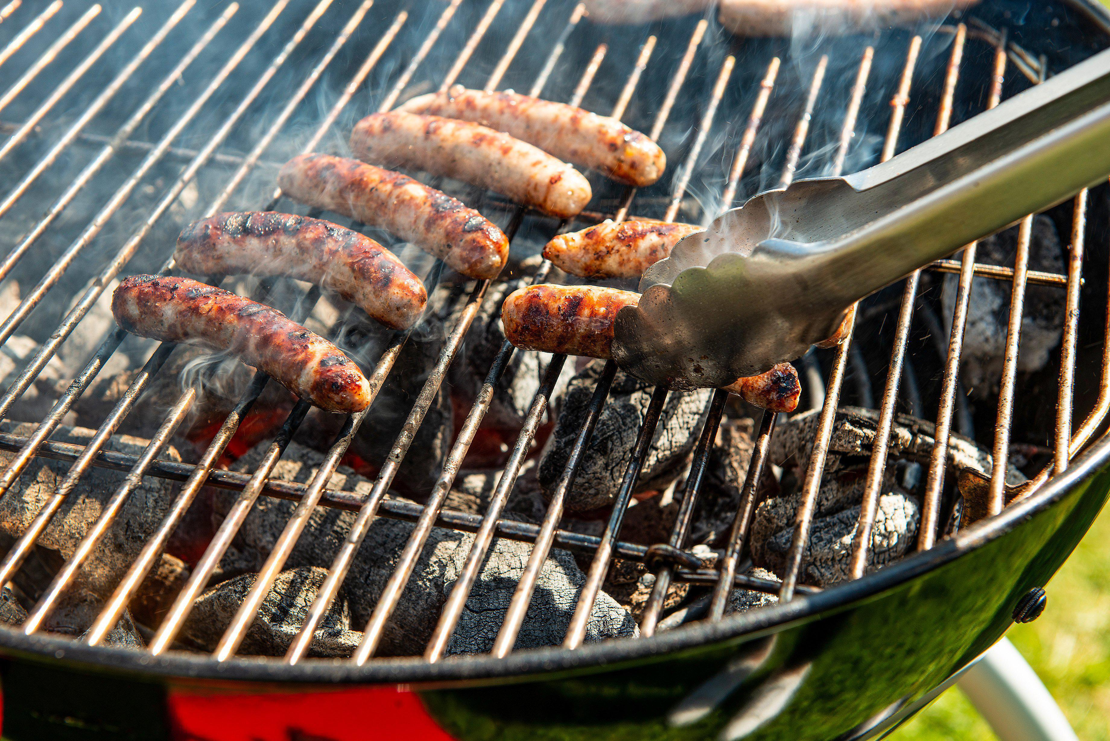 Würstchen auf einem Grill, eine Zange hält eine.