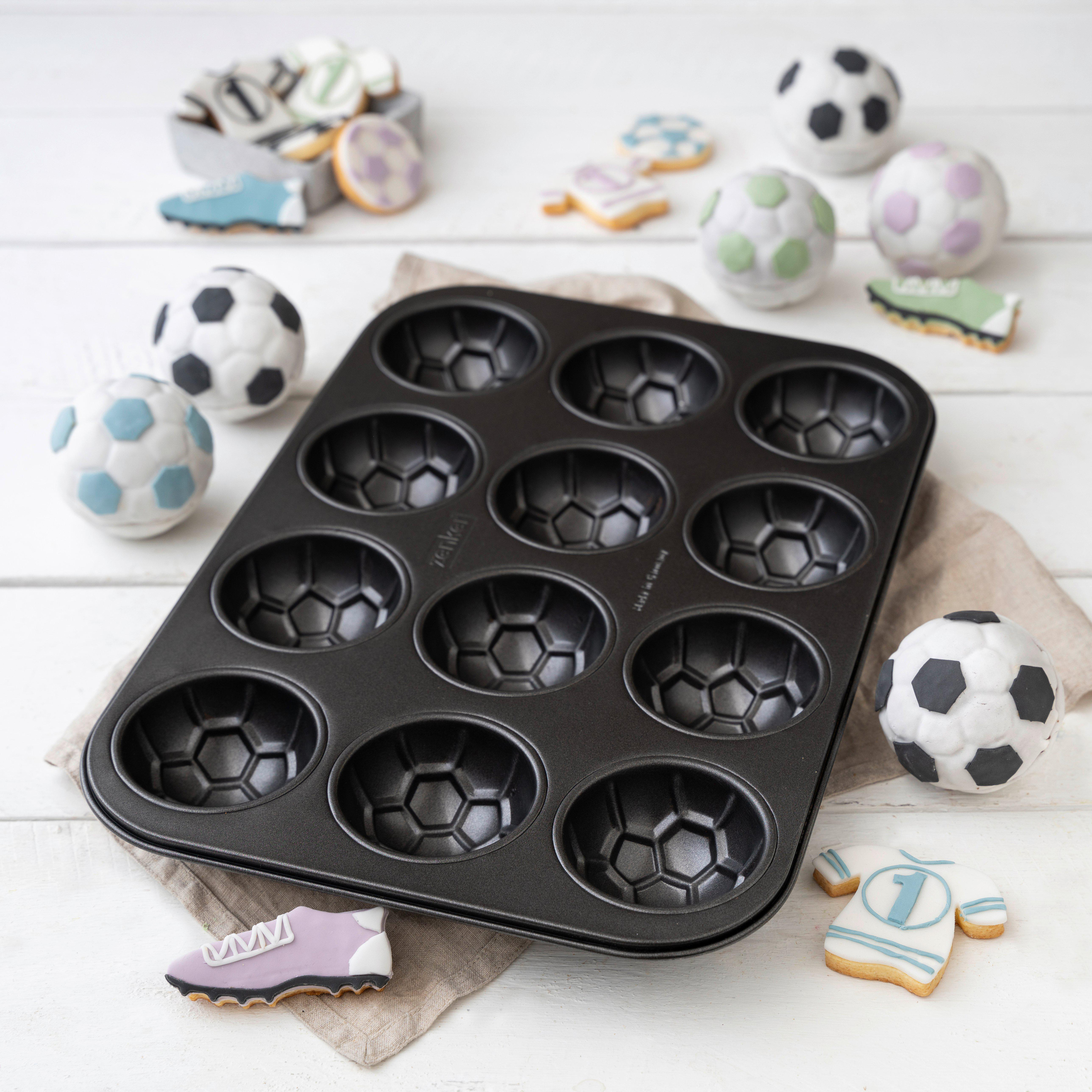 Eine schwarze Muffinbackform im Fußballmuster auf weißem Tisch, umgeben von Fußball-Keksen und runden Küchlein.