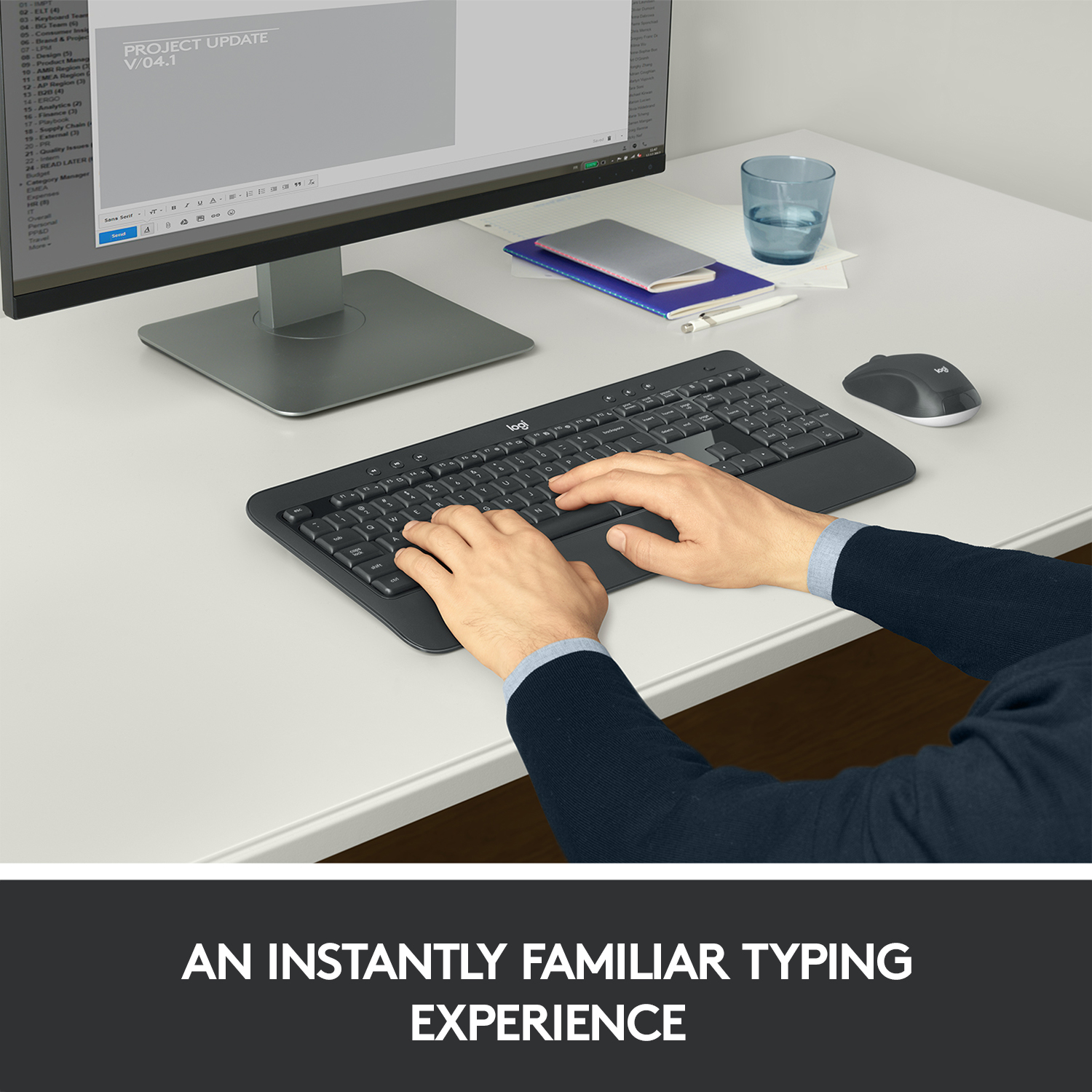Hände tippen auf einer schwarzen Tastatur. Ein Monitor zeigt Text an. Maus und blauer Becher auf dem Schreibtisch.