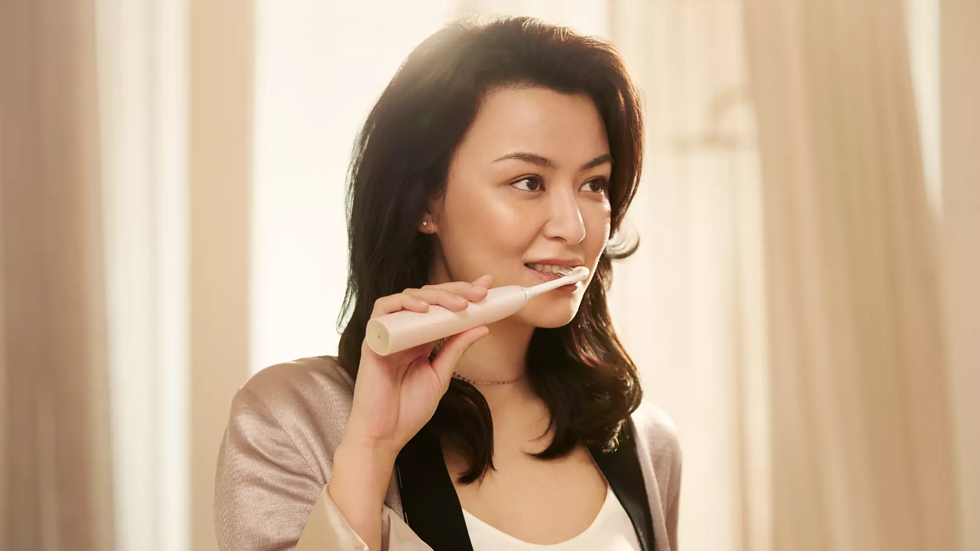 A woman brushing her teeth with an electric toothbrush, smiling, sunlit background.