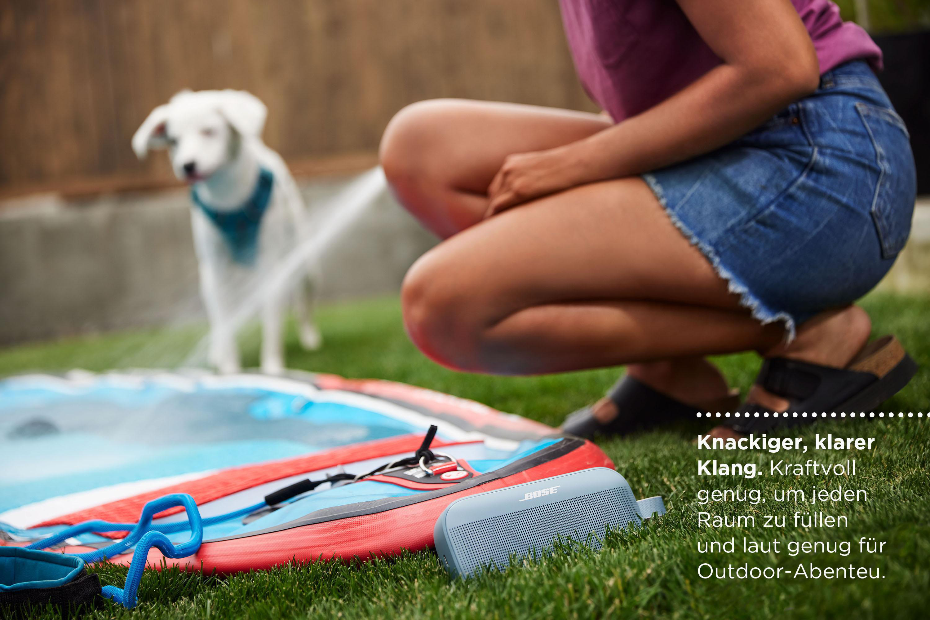Frau und Hund mit Lautsprecher und Windsurfbrett auf Gras.