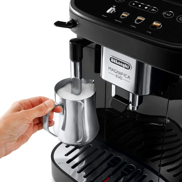 A person's hand frothing milk into a metal pitcher, next to a coffee machine.