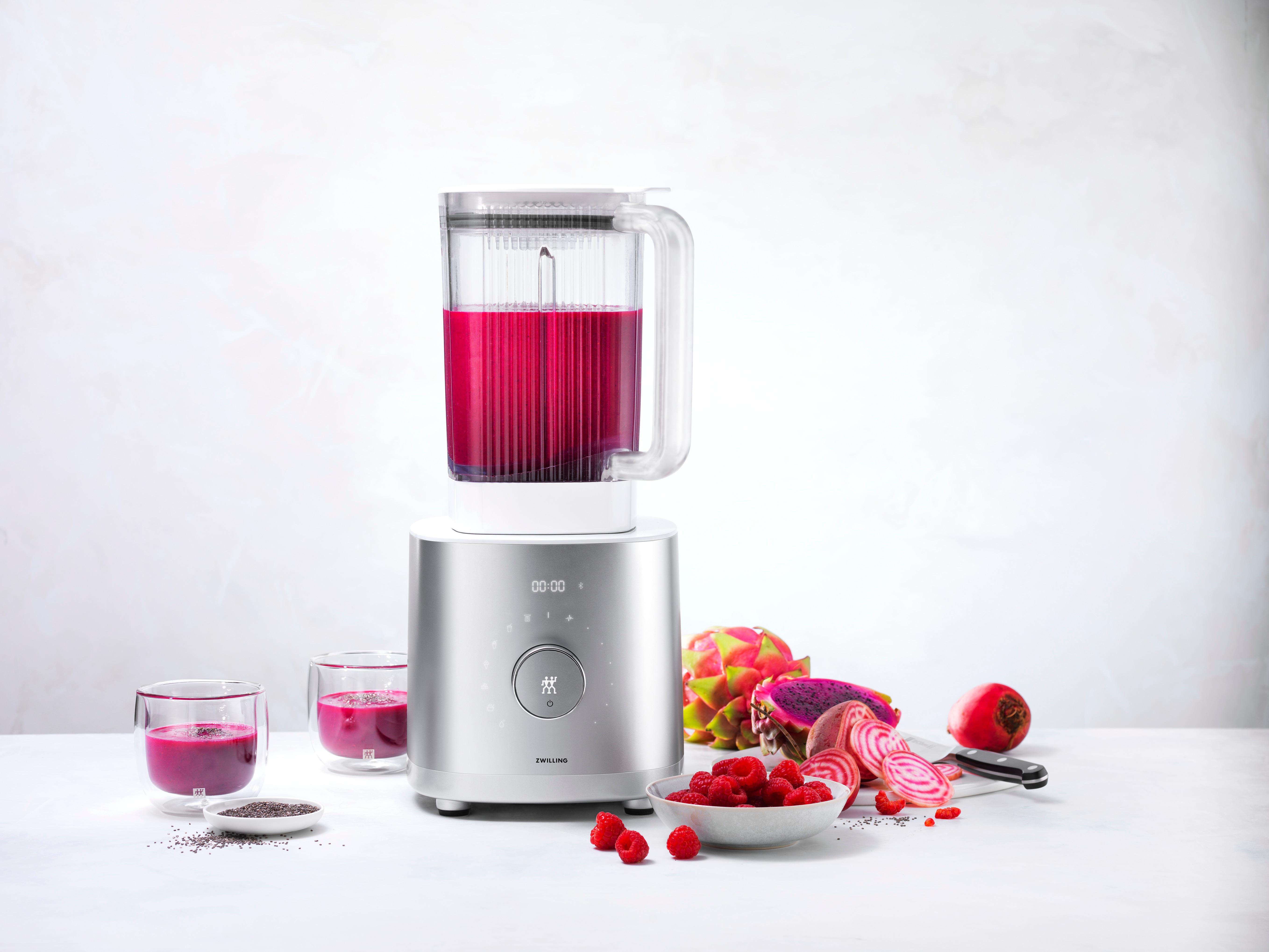 Mixer mit lila Flüssigkeit, Drachenfrucht und Himbeeren auf weißer Oberfläche.