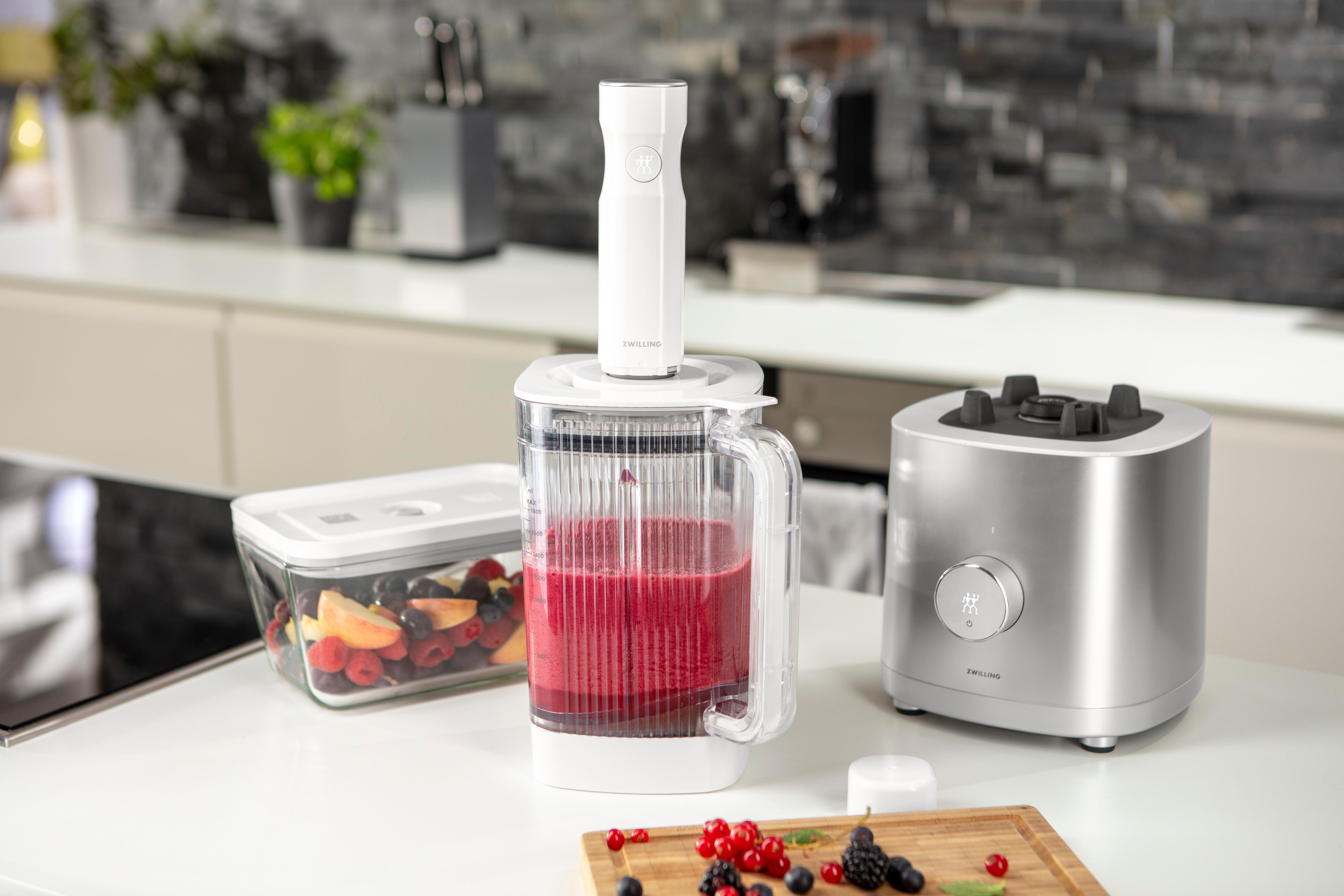 Mixer mit roter Flüssigkeit und Früchten. Ein Behälter mit Beeren und andere Küchenartikel sind auf der Theke.