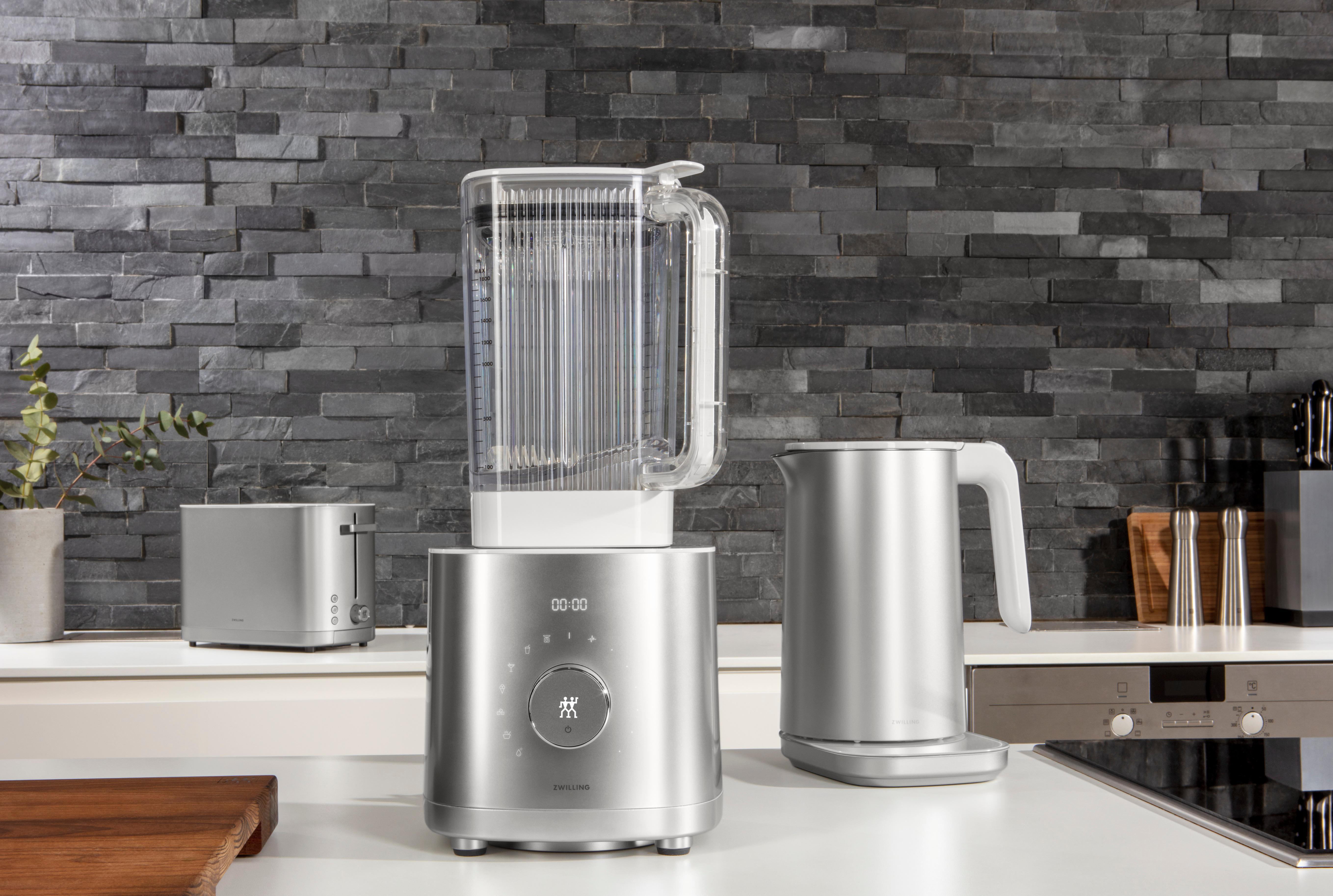 Silberne Küchengeräte auf einer Arbeitsplatte vor einer grauen Ziegelmauer. Mixer, Toaster, Wasserkocher.