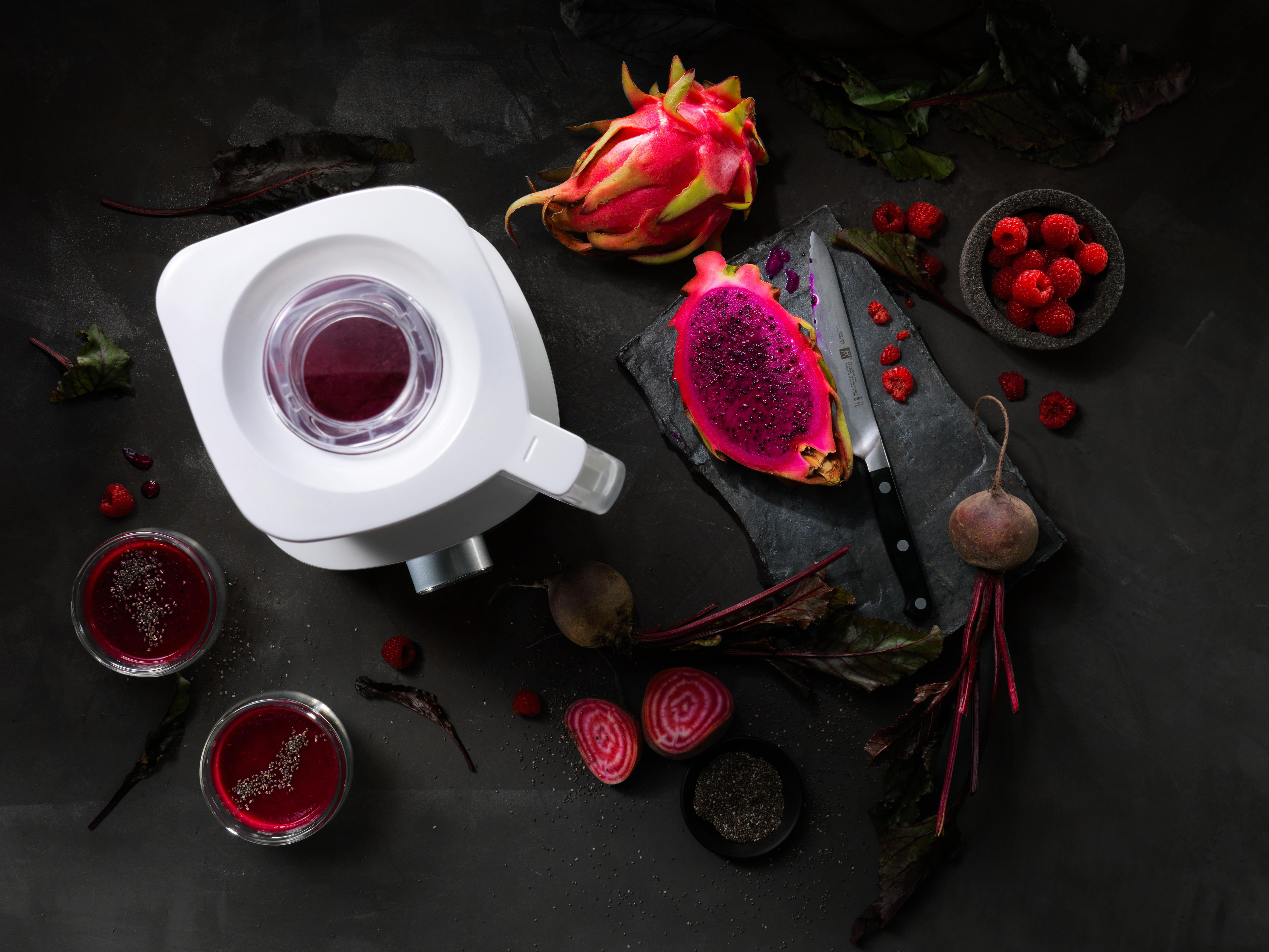 Draufsicht auf einen Entsafter mit geschnittener Drachenfrucht, Rüben und Himbeeren auf schwarzer Oberfläche.
