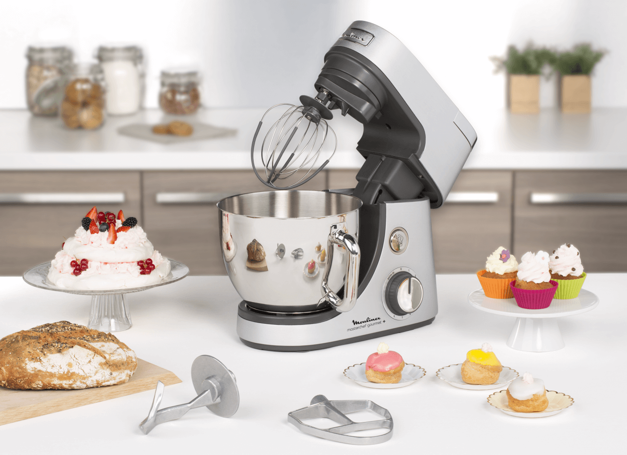 Silver and white stand mixer with accessories and baked goods on a white counter.