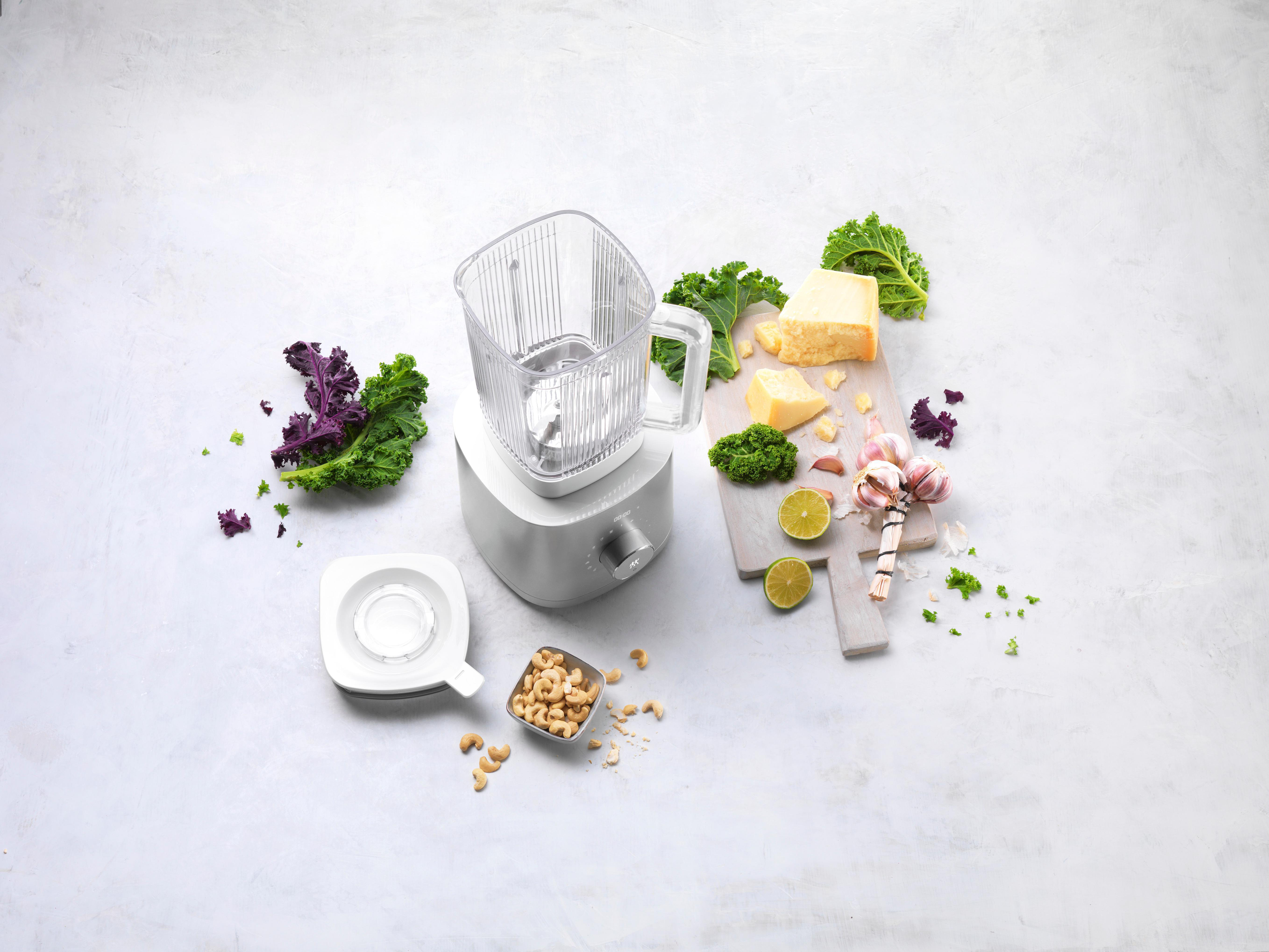 Ein Mixer mit Zutaten wie Grünkohl, Käse und Cashewnüssen auf heller Fläche.