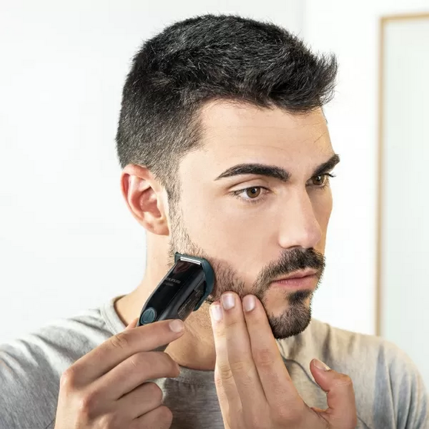 Hombre se afeita la barba con recortadora. Sostiene la piel. El fondo es una pared blanca.