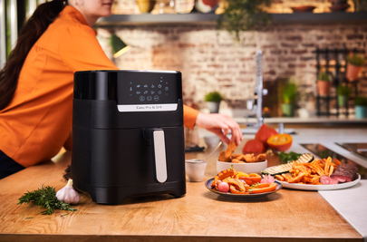 An air fryer with food on a wooden countertop. A person is in the background.