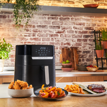 An air fryer with fried foods and a steak on a wooden surface, against a brick wall.