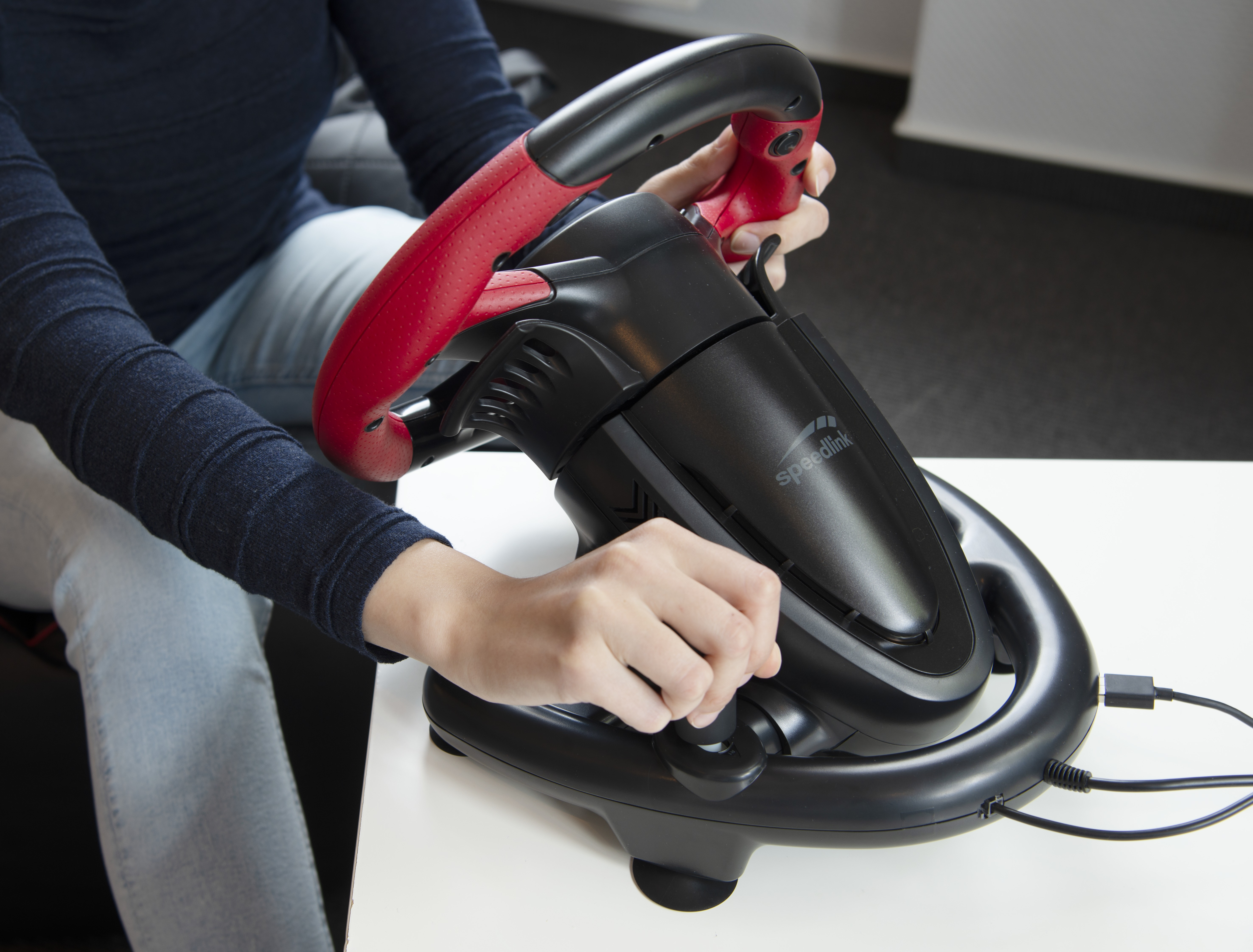 Person hält ein schwarz-rotes Rennlenkrad. Die Marke Speedlink ist sichtbar.