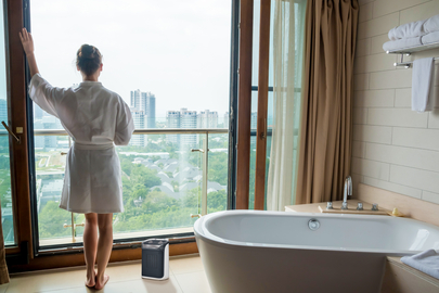 Woman in white robe stands by a window, looking at city, a bathtub to the right, and a heater.