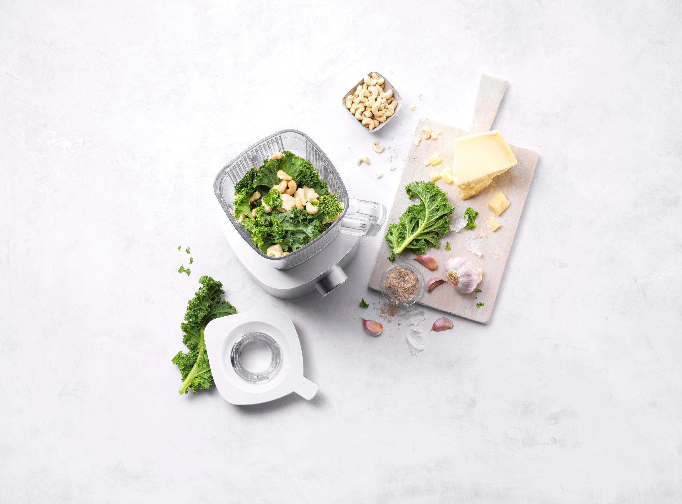 Mixer mit Grünkohl und Cashewnüssen. Käse und Knoblauch auf einem Holzbrett.