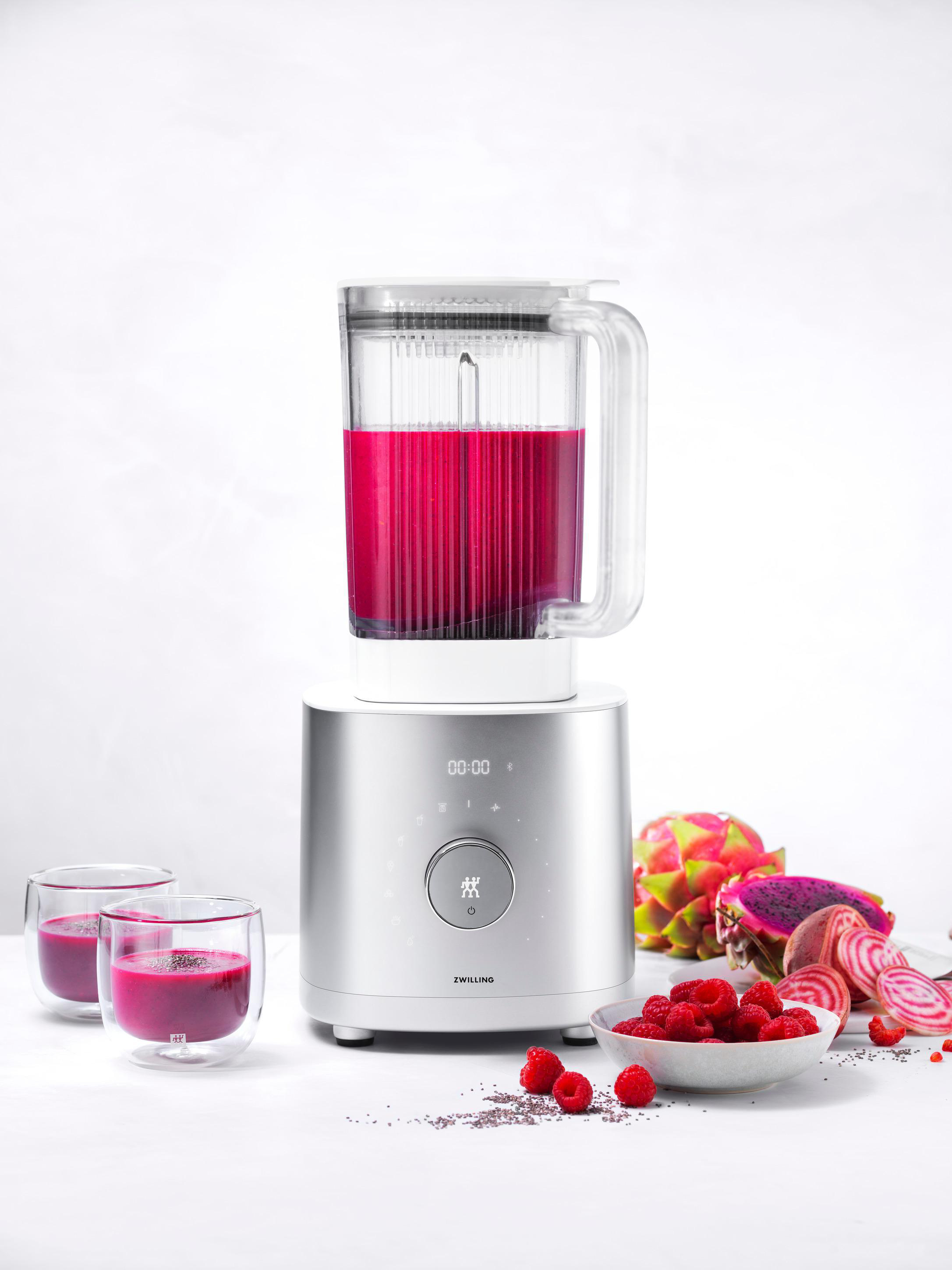 Mixer mit rosa Flüssigkeit, Himbeeren, Drachenfrucht und Rote Bete auf weißer Oberfläche.