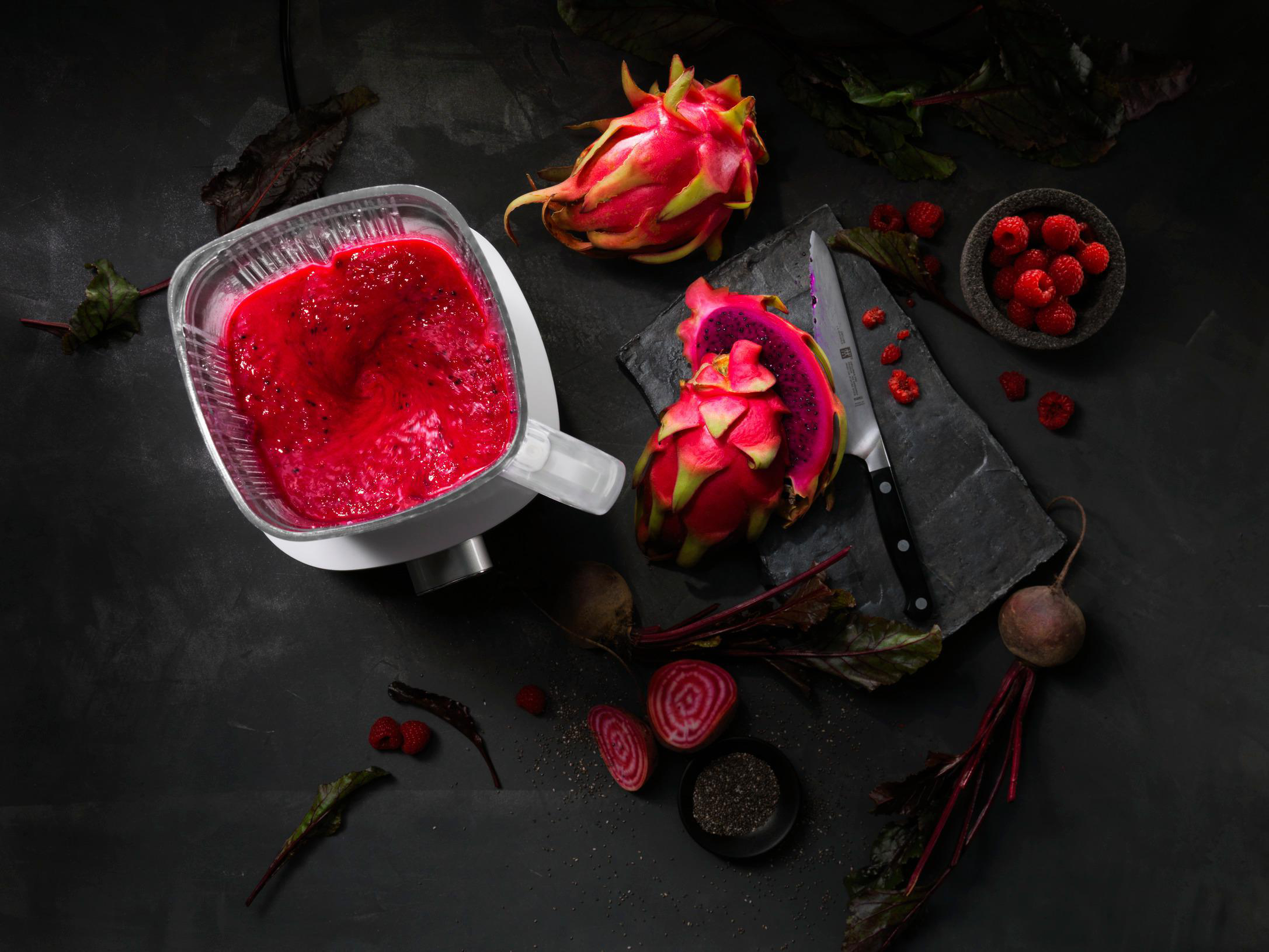 Mixer mit rotem Smoothie, Drachenfrucht, Himbeeren und Messer auf dunkler Oberfläche. Draufsicht.