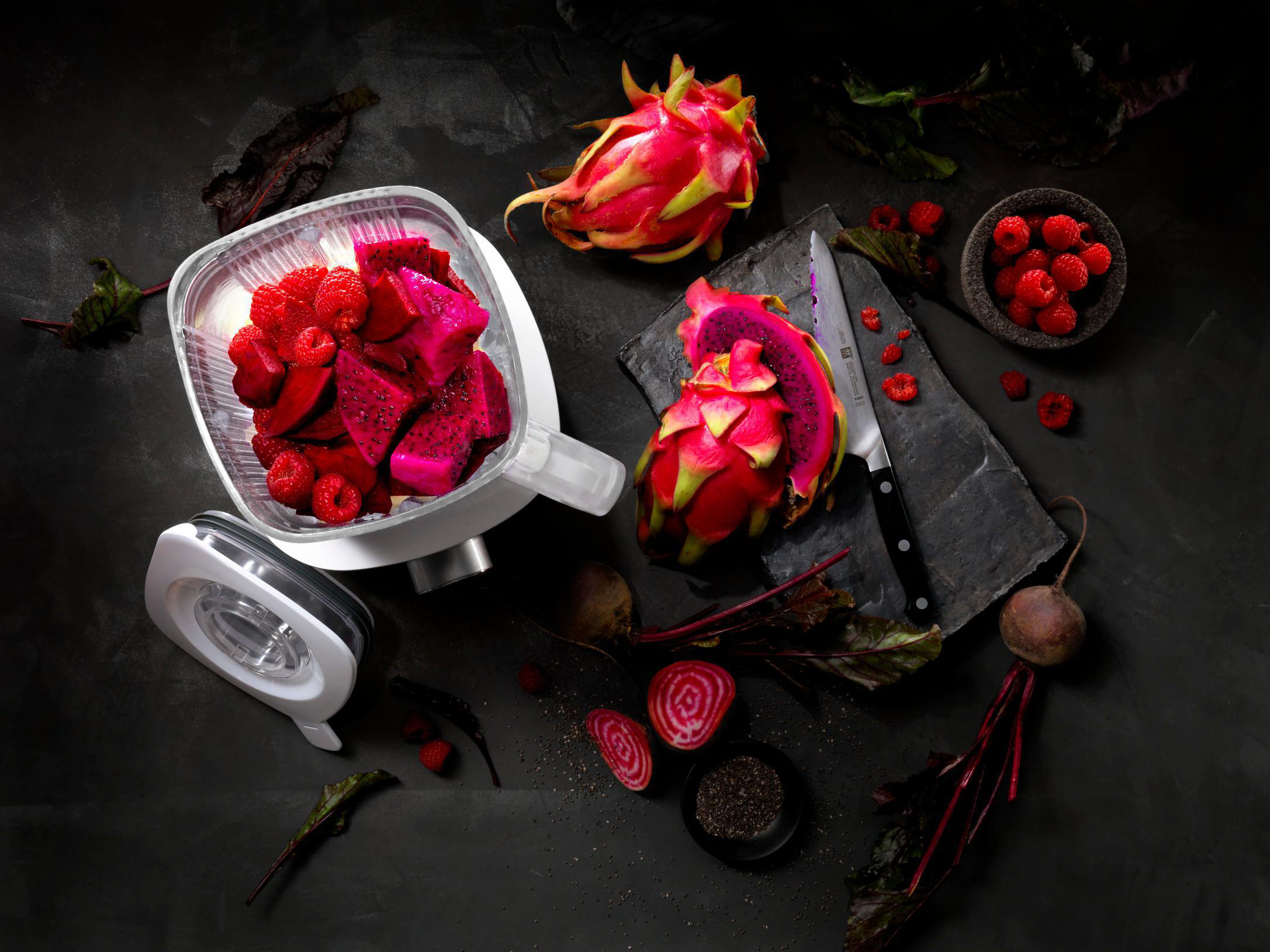 Ein Mixer mit geschnittener Drachenfrucht und Himbeeren, mit Messer und Rüben. Schwarzer Hintergrund.