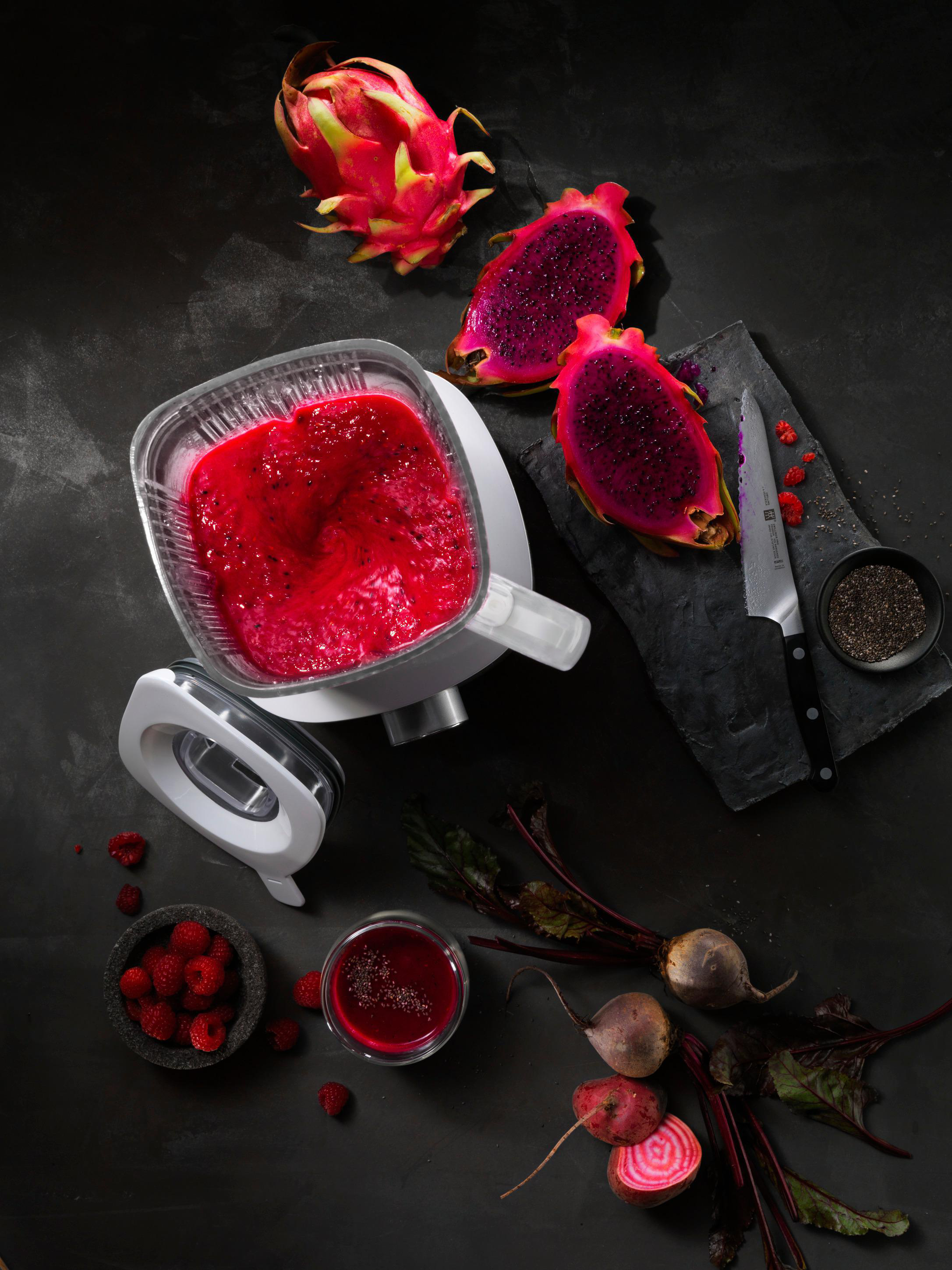 Draufsicht auf einen Mixer mit roter Flüssigkeit und Früchten, darunter Drachenfrucht, Himbeeren und Rüben.