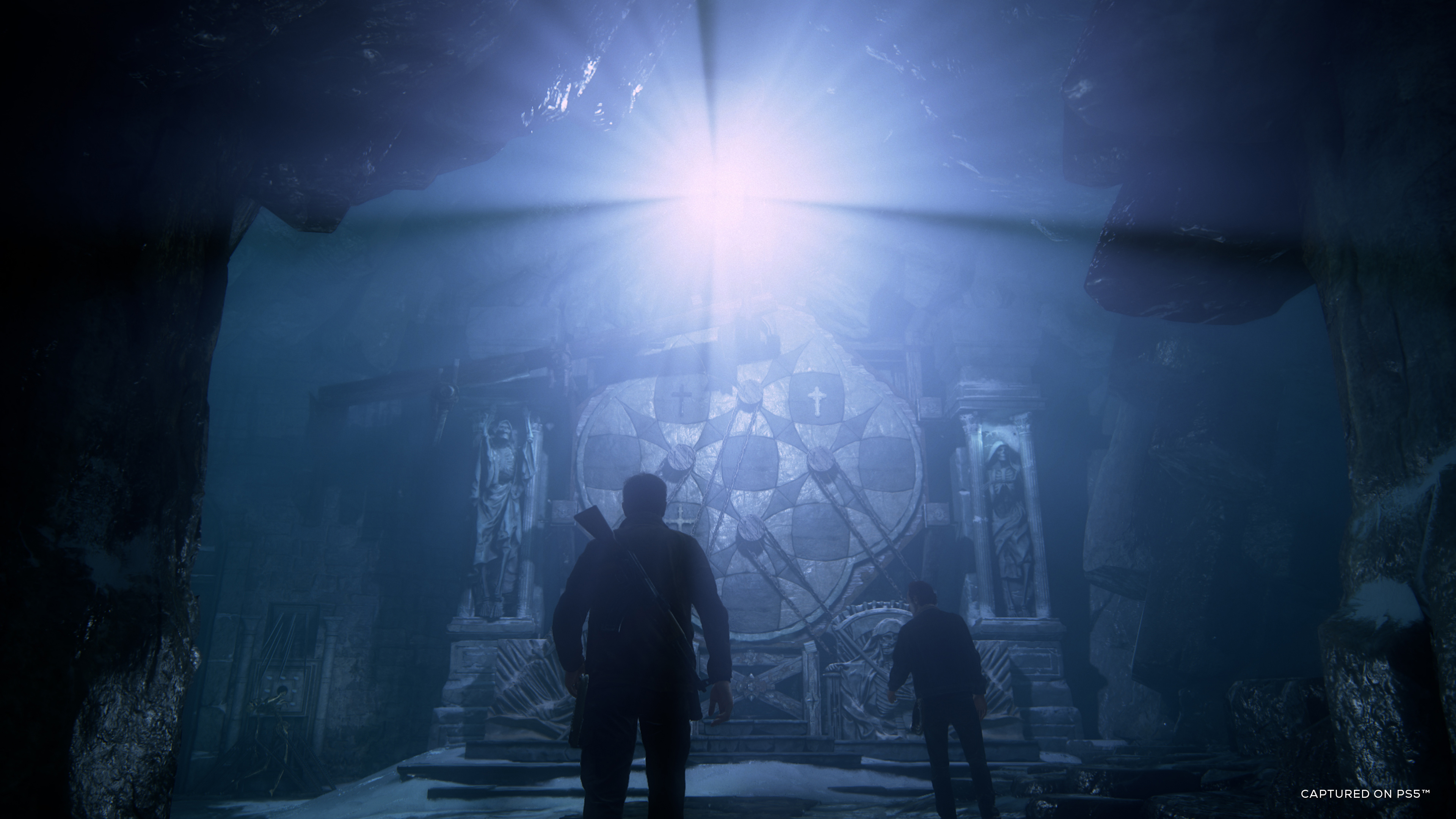 Zwei Figuren in einer Höhle, beleuchtet von einer hellen Lichtquelle. Steinstruktur im Hintergrund.