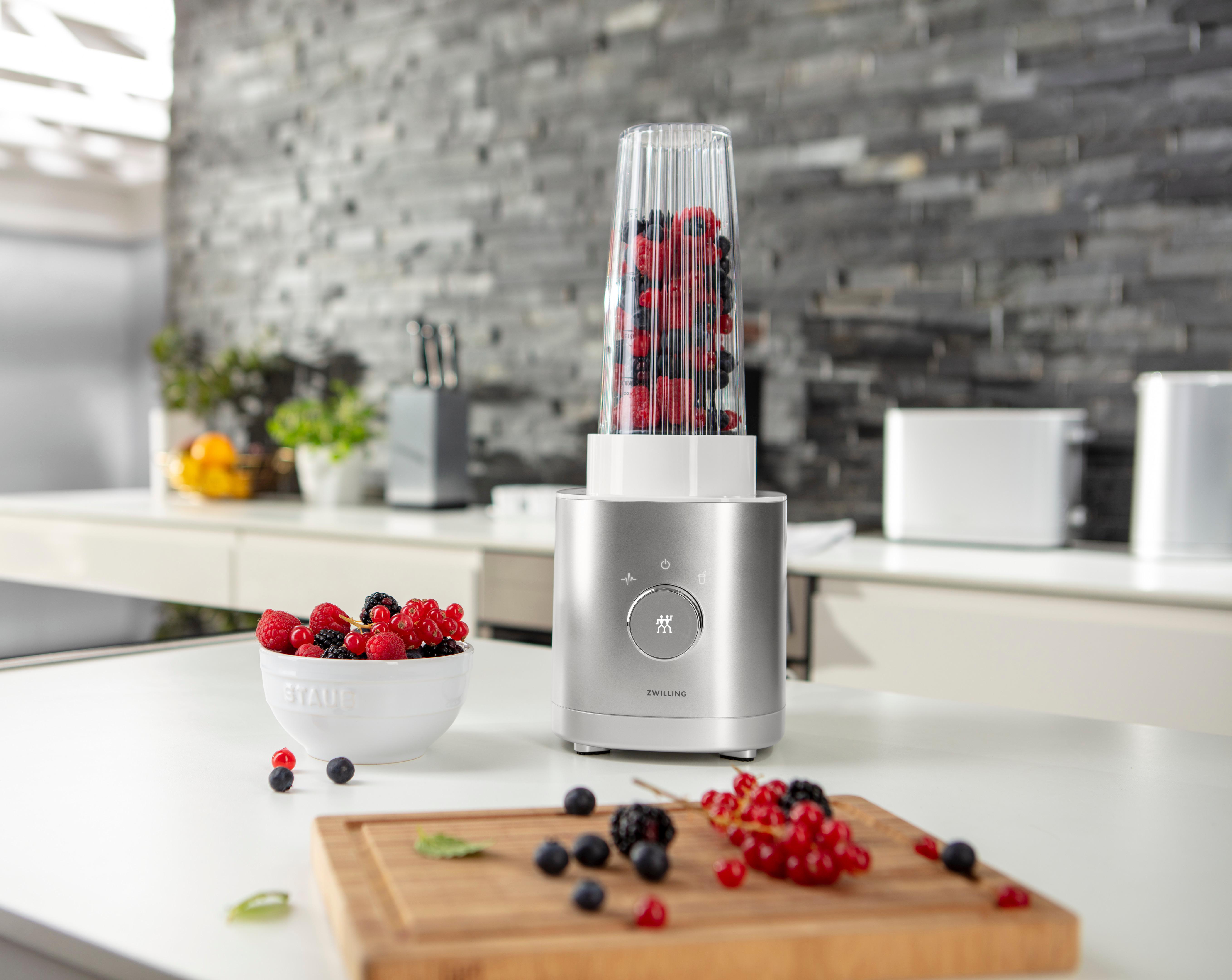 Mixer mit gemischten Beeren auf weißer Theke, mit Schüssel und Schneidebrett.
