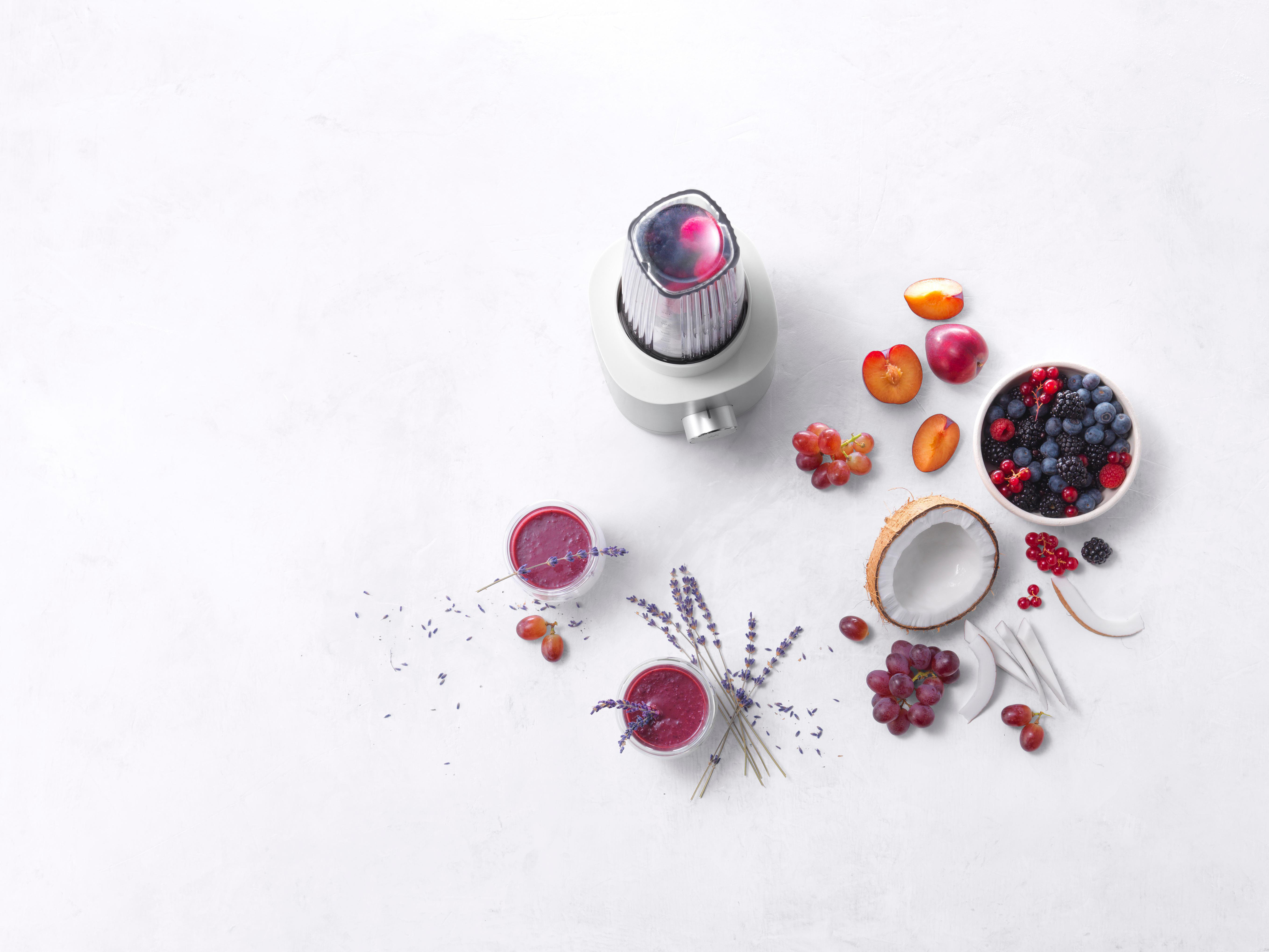 Draufsicht auf Mixer, Früchte und zwei Smoothies auf weißer Oberfläche.