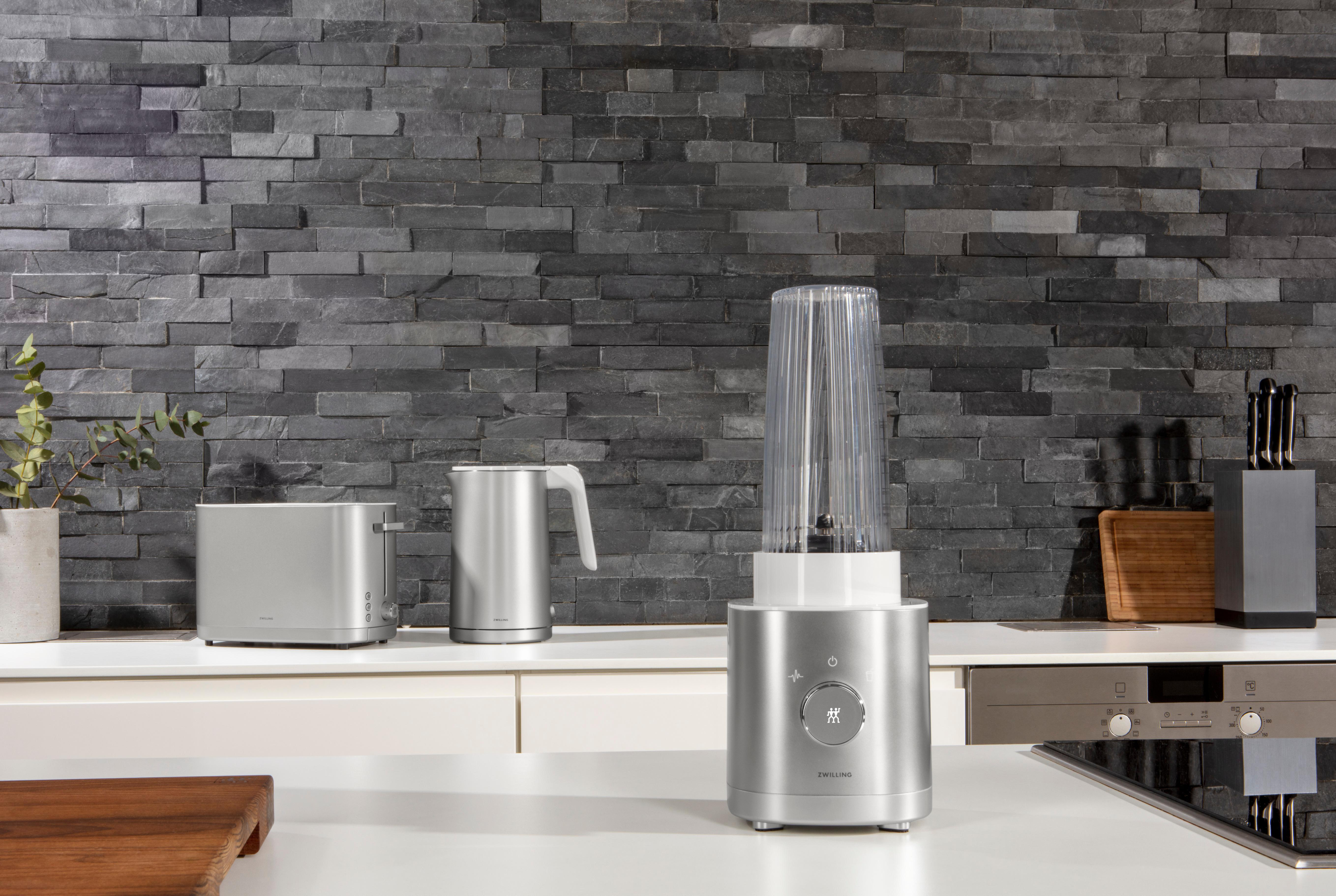 Silberne Küchengeräte: Mixer, Toaster, Wasserkocher und Messer vor Steinwand-Hintergrund.