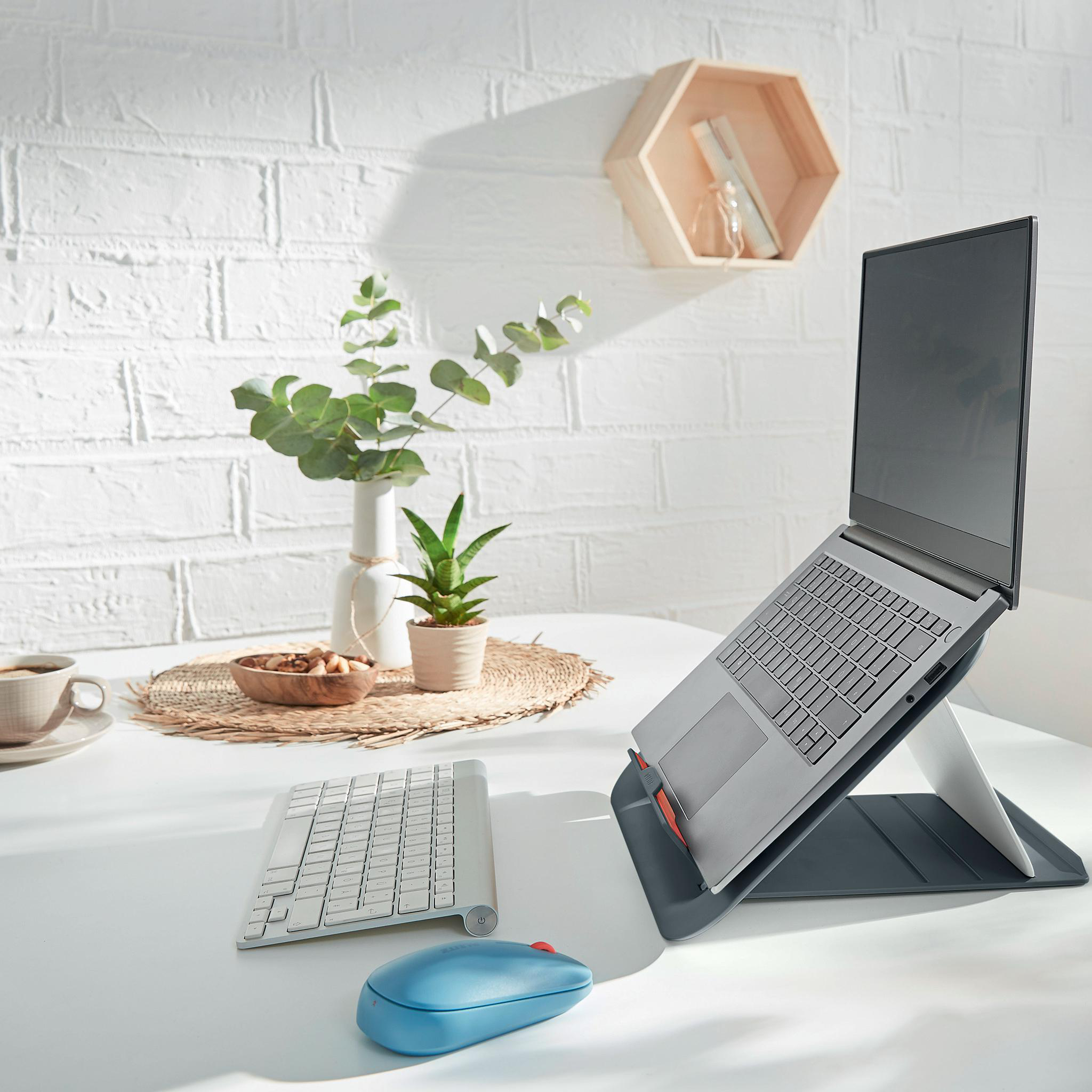 Laptop auf Ständer mit Tastatur und Maus auf weißem Tisch. Dekorative Elemente im Hintergrund.
