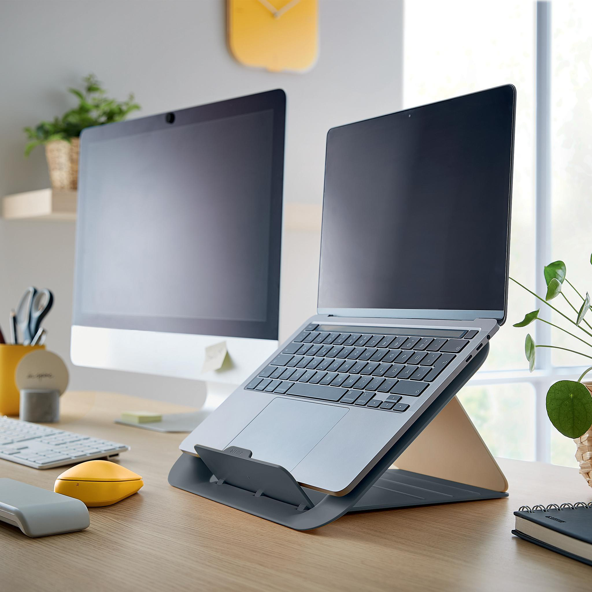 Laptop auf Ständer, Computer und Pflanze auf einem Holzschreibtisch. Gelbe Uhr an der Wand.