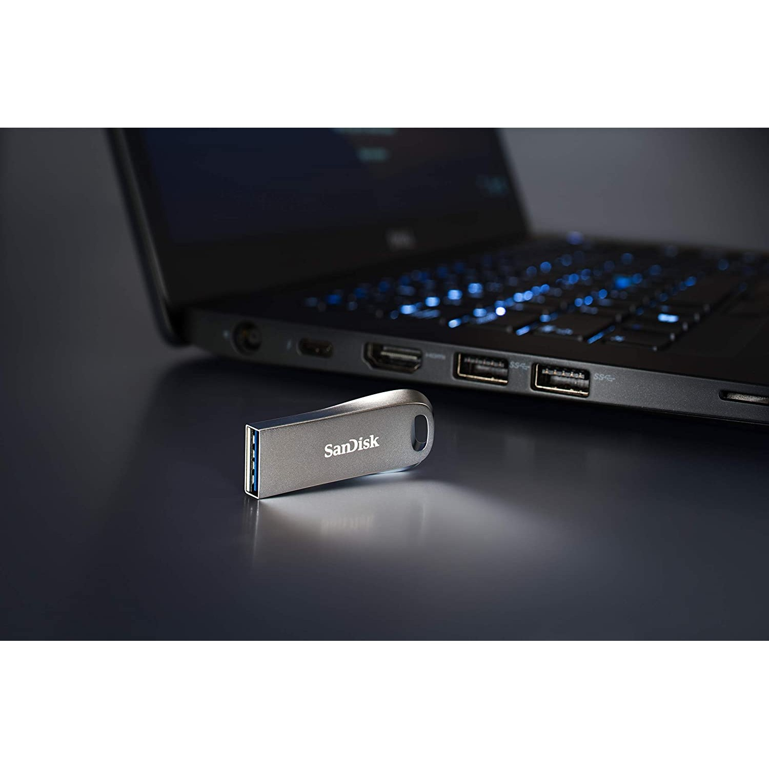 Unidad USB SanDisk plateada junto a una computadora portátil con teclado brillante, sobre una superficie oscura.