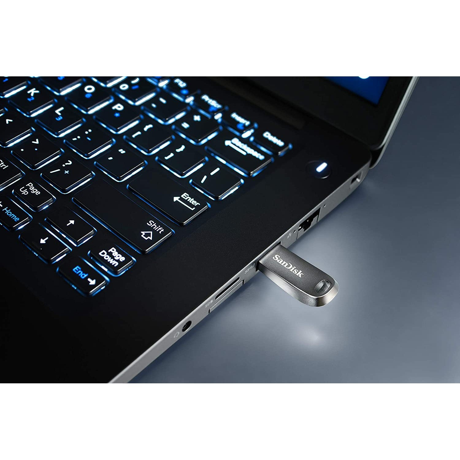 Portátil con teclado iluminado, una unidad USB plateada conectada. Fondo borroso.